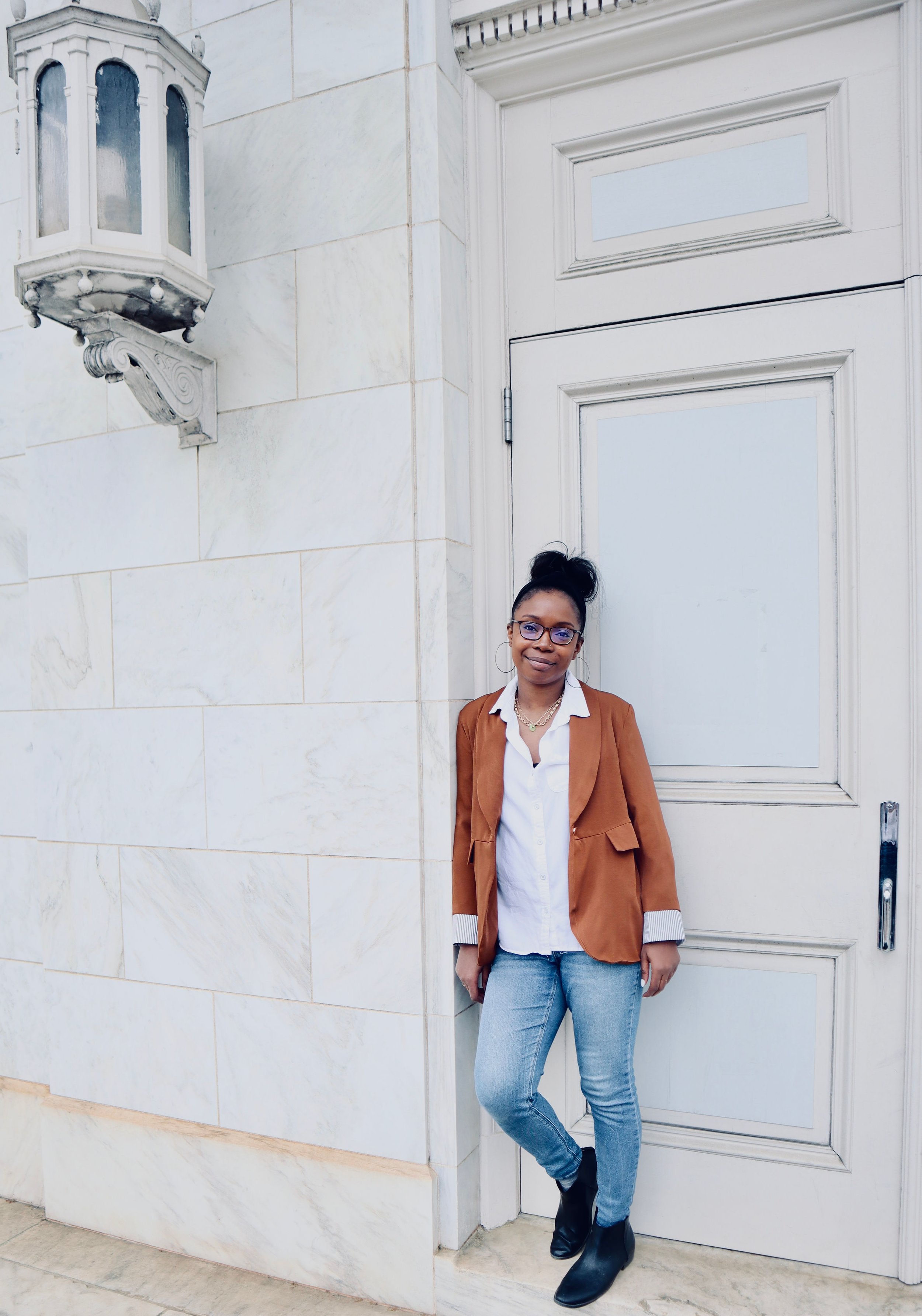 Erica, a black female wearing glasses and hair in a top bun, leans against a doorframe outside. She wears blue jeans, black boots, a whit button down and brown blazer.