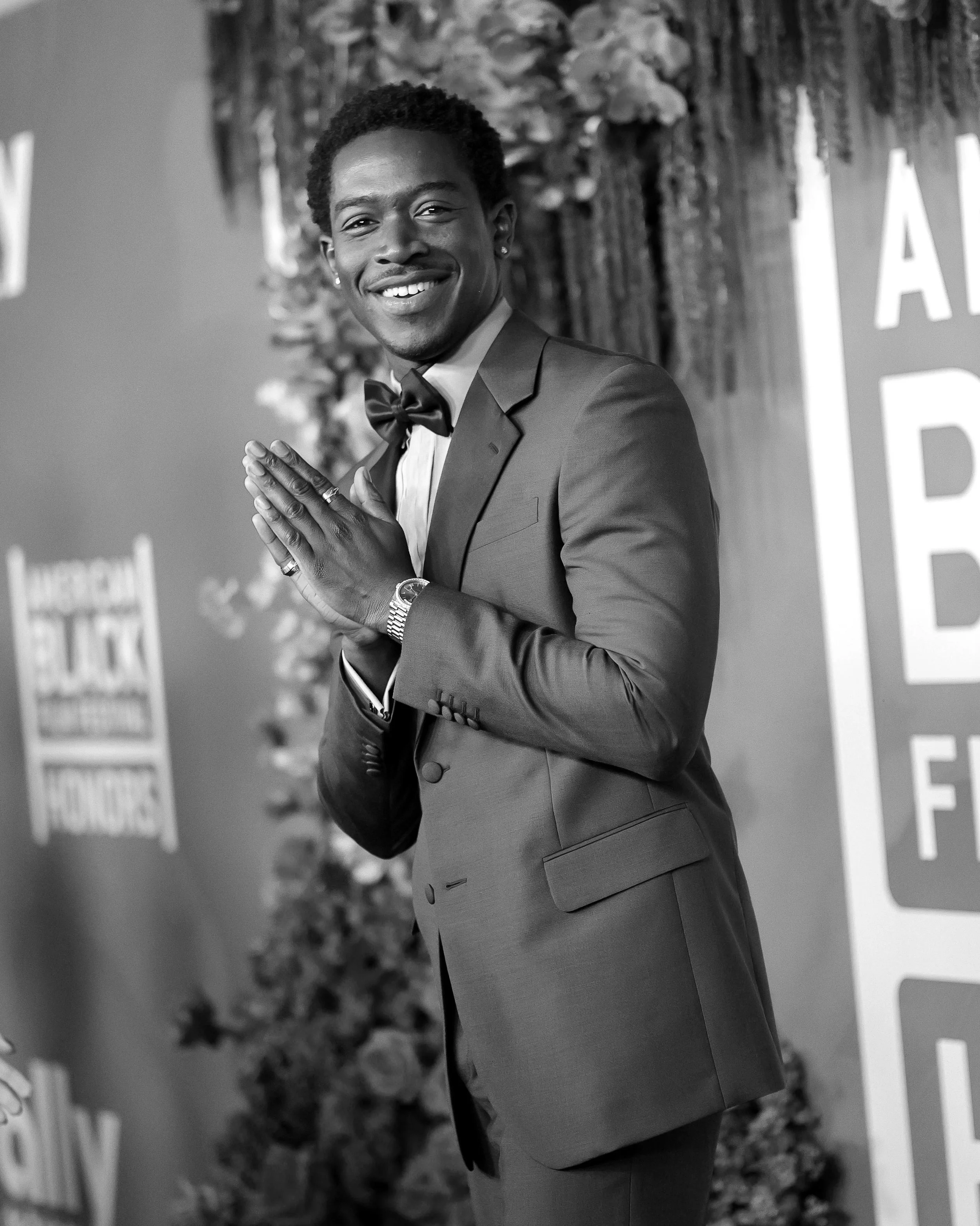 A smiling man dressed in a suit with a bow tie, standing at an event, holding his hands together in a respectful gesture, with a decorated background and partial text visible.