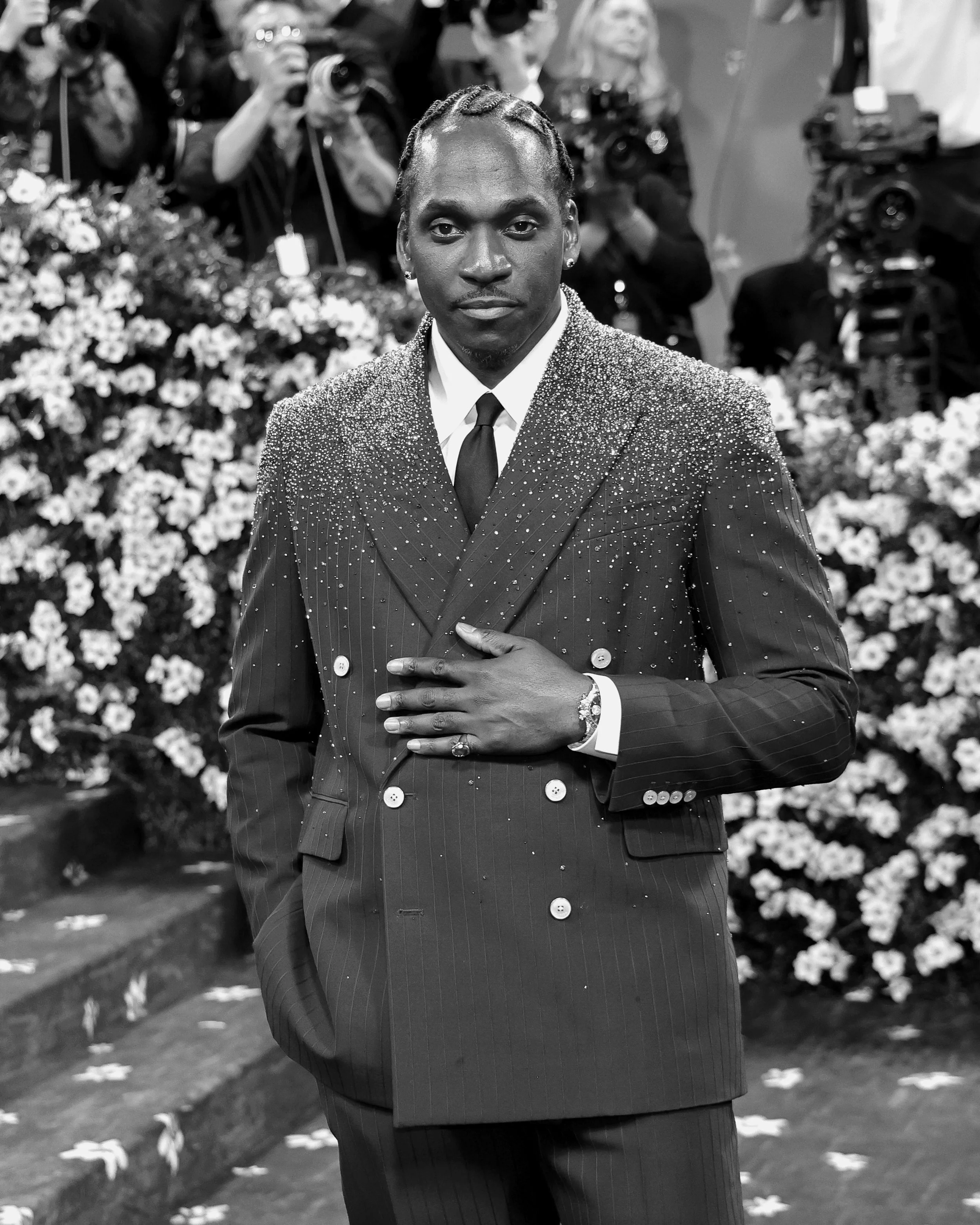 A man dressed in a pinstripe suit with decorative embellishments, standing with one hand on his chest at a formal event, with photographers and floral decorations in the background.