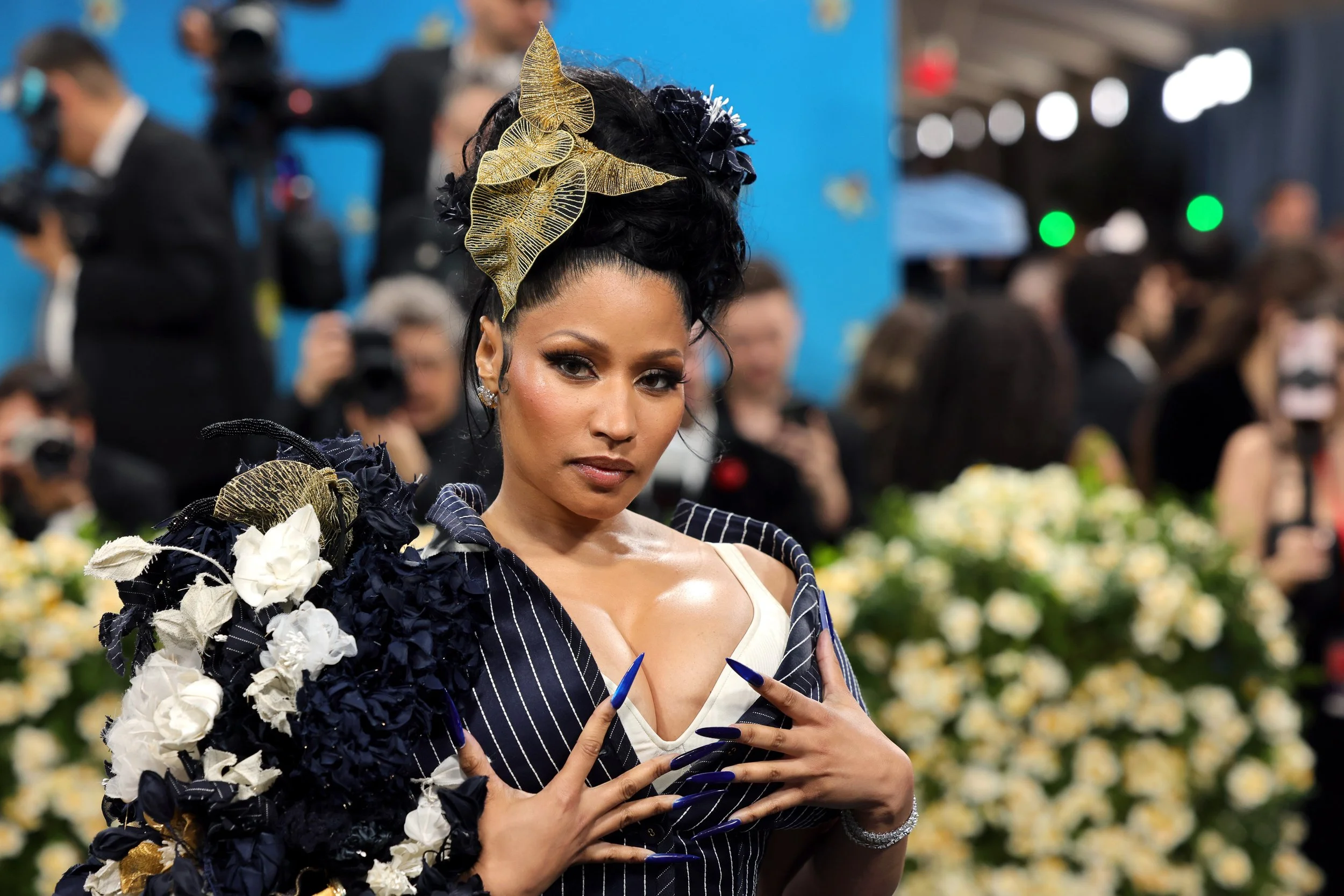 A woman with dark hair adorned with gold leaves, wearing a black and white striped outfit with floral embellishments, poses at a formal event surrounded by photographers and other attendees.