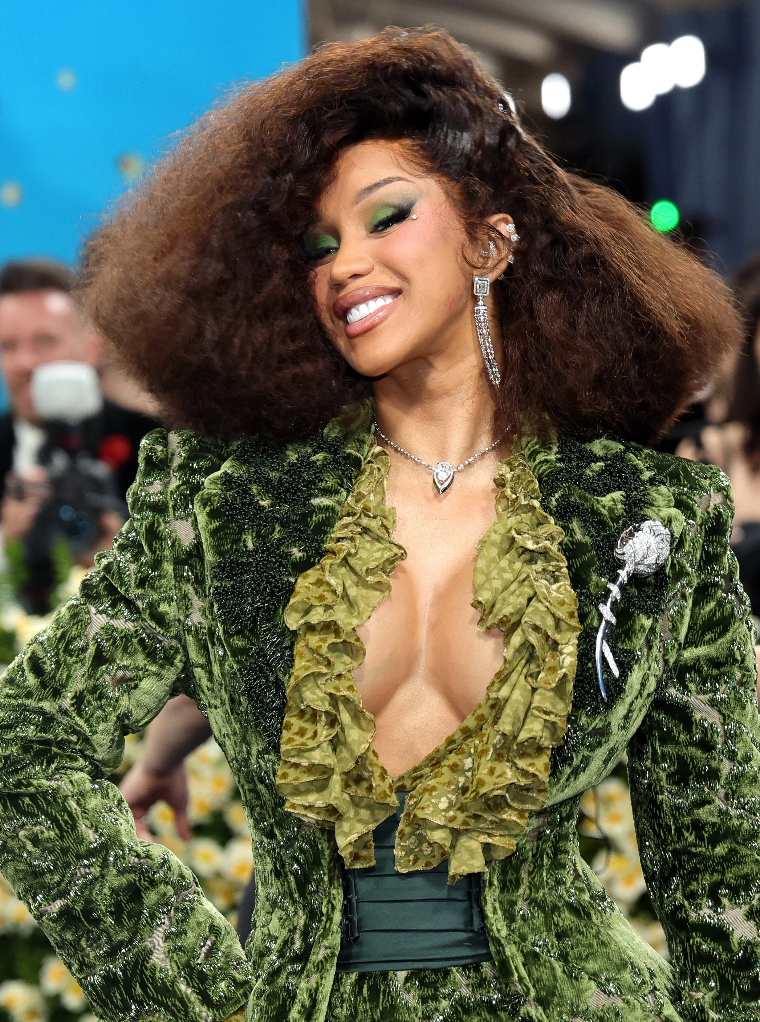 A woman with curly brown hair smiling, wearing a green velvet jacket with ruffled neckline, and adorned with jewelry, including earrings and a necklace, at a public event.