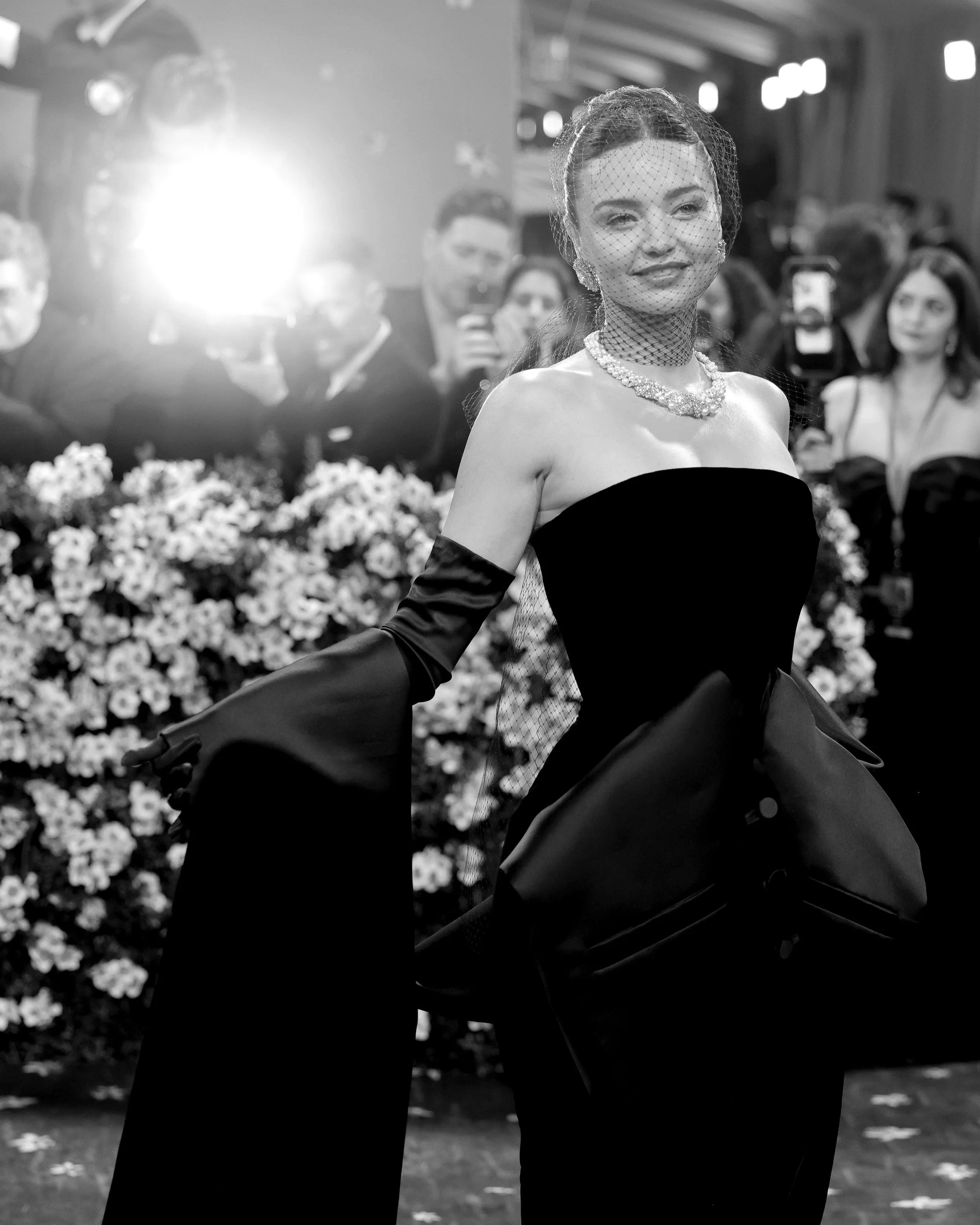 A woman in a black strapless gown with long gloves and diamond jewelry, smiling, surrounded by photographers and crowd at a formal event.
