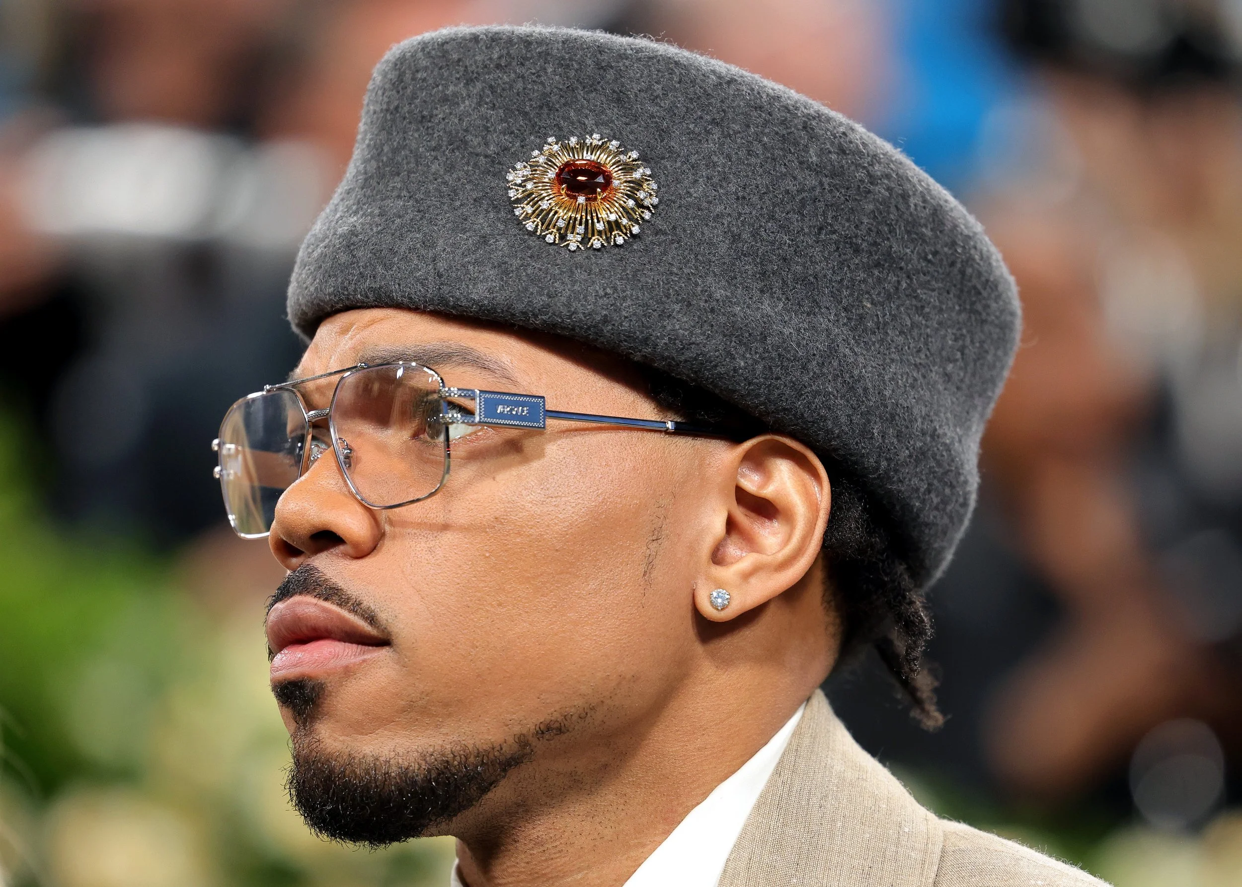 Close-up of a man wearing a gray beret with a decorative pin, glasses, diamond earrings, and a suit, at a public event.