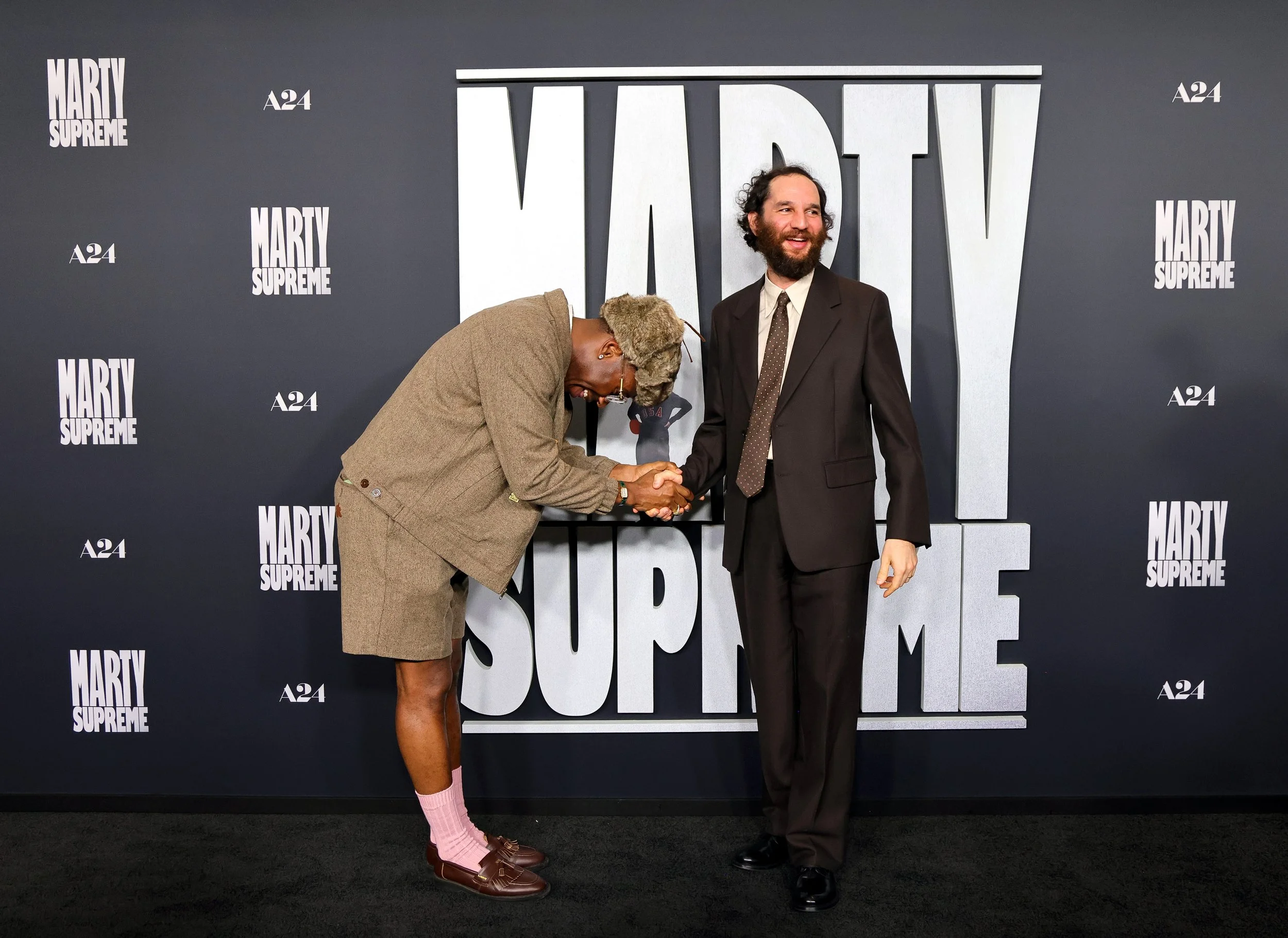 Two men shaking hands in front of a backdrop with the text 'MARTY SUPREME' and 'A24'. One man is dressed in a suit, the other in a beige jacket, shorts, pink socks, and brown shoes.