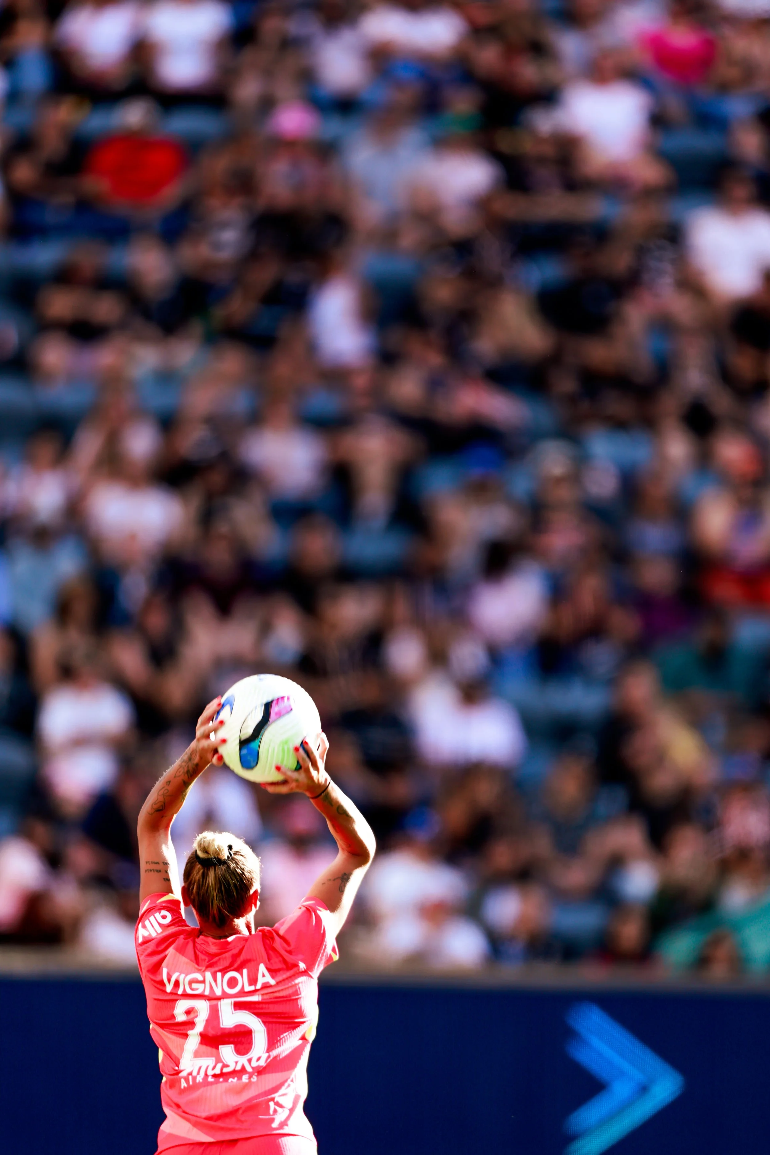 Female soccer player wearing a pink jersey with the number 25 and the name Vignola, holding a white soccer ball with colorful markings during a game with a large audience in the background.