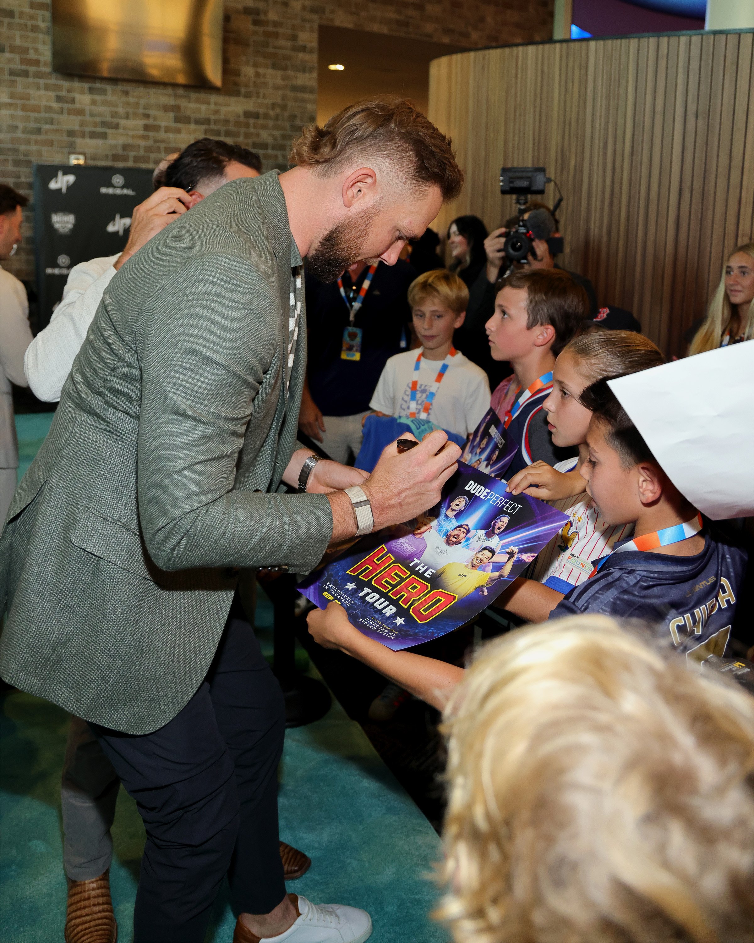A man signing autographs for children at a signing event, with children holding posters of a soccer player and wearing medals, in an indoor setting with a brick wall and photographers in the background.