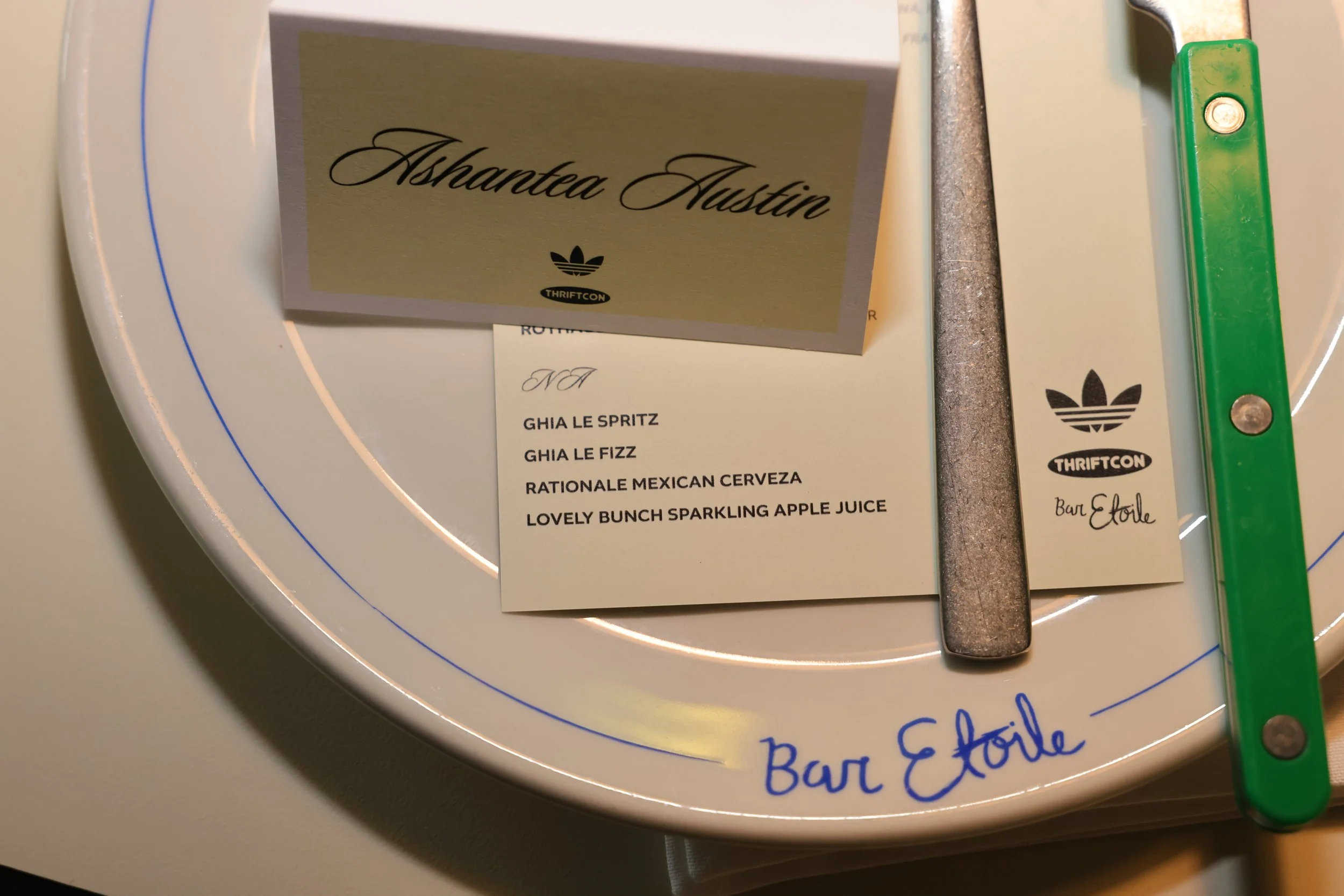 A circular white tray with blue accents labeled 'Bar Etoile', featuring a name card with 'Ashante Austin' and 'ThriftCon' logo, a menu reading 'N/A' with items including 'Ghia Le Spritz', 'Ghia Le Fizz', 'Rationale Mexican Cerveza', and 'Lovely Bunch