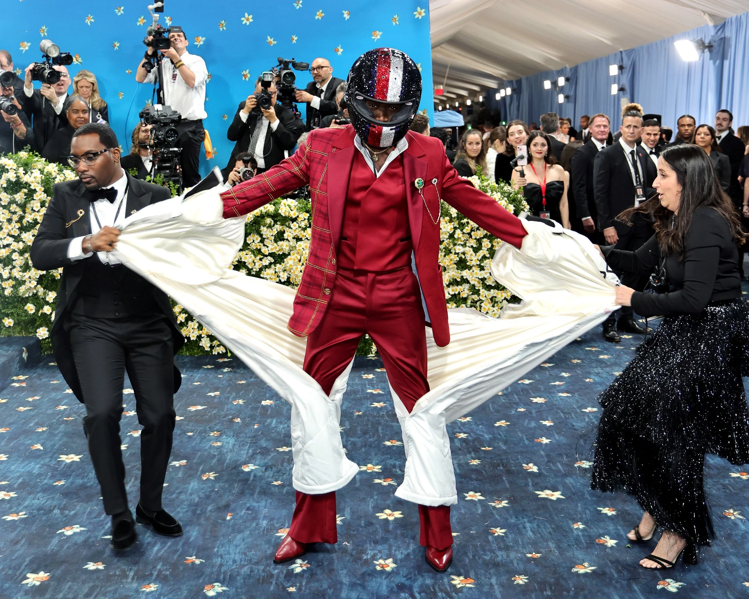 A person in a red suit and a shiny helmet is holding a white fabric with two assistants, one in a black tuxedo and the other in a black dress with sparkles. There are photographers and guests in the background at a formal event.
