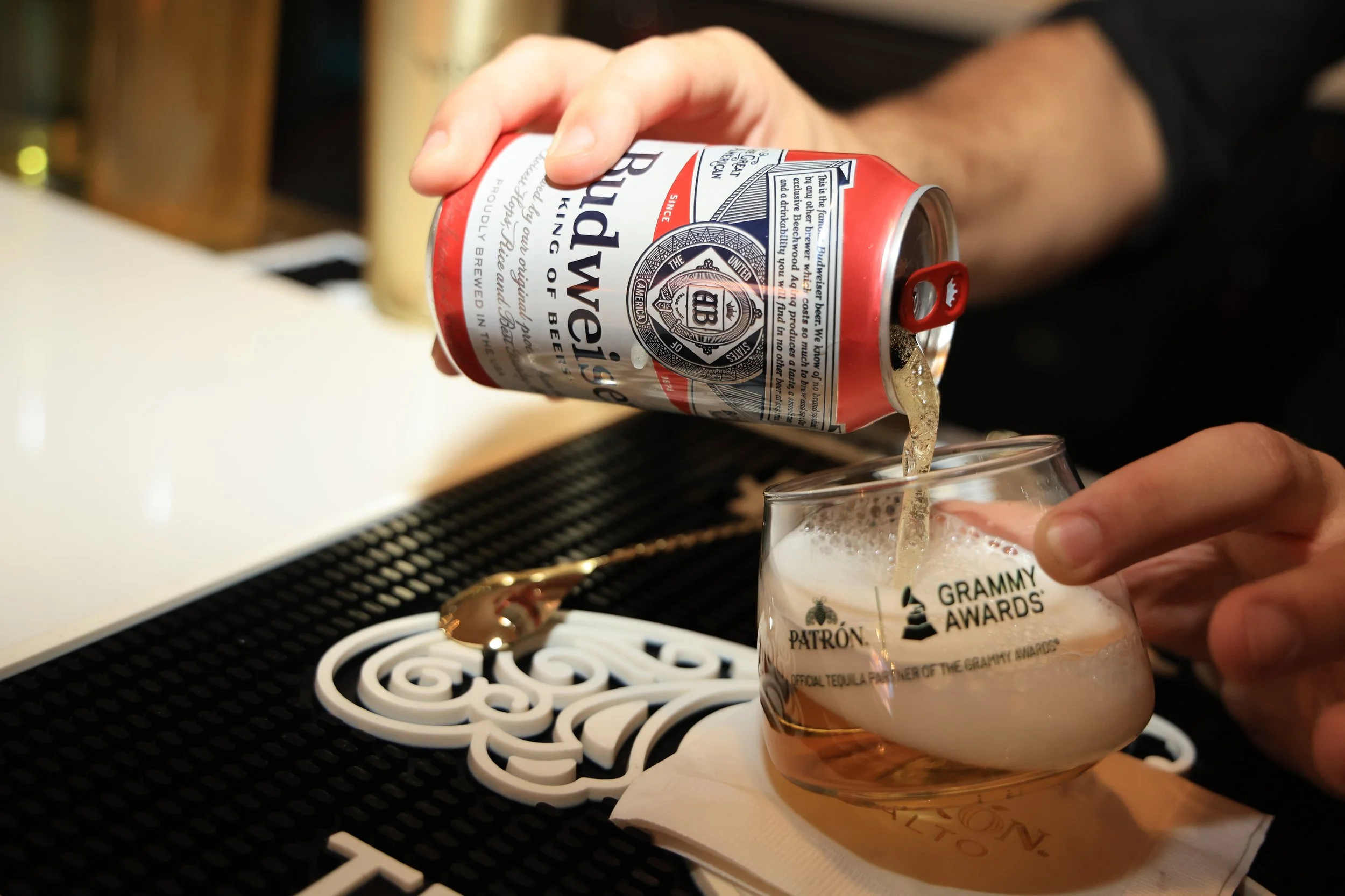 A person pouring Budweiser beer into a glass with the Grammy Awards logo on it, sitting on a bar counter.
