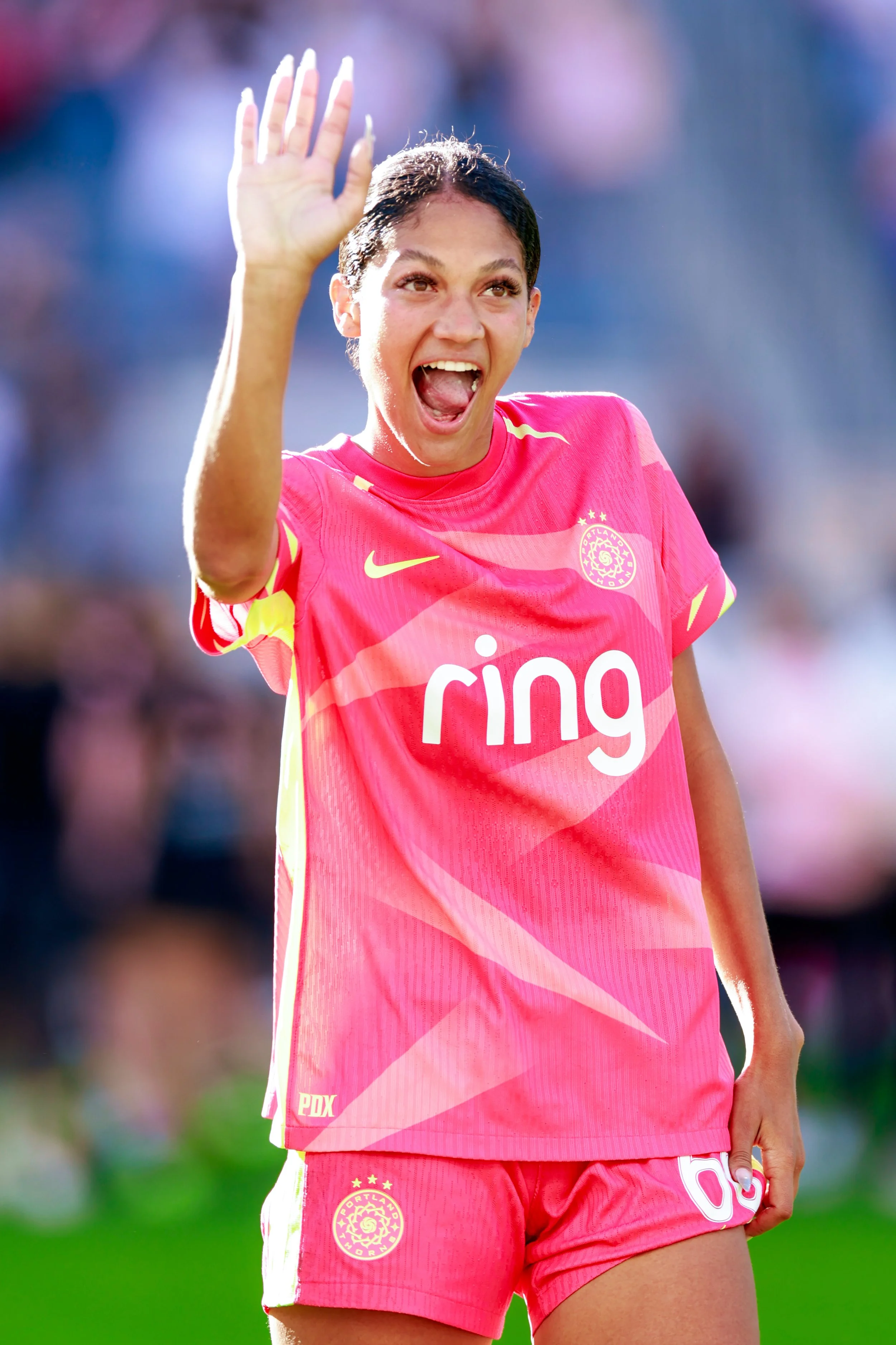A female soccer player waving and celebrating on the field, wearing a pink team jersey and shorts with a logo that reads 'ring' and the number 65.