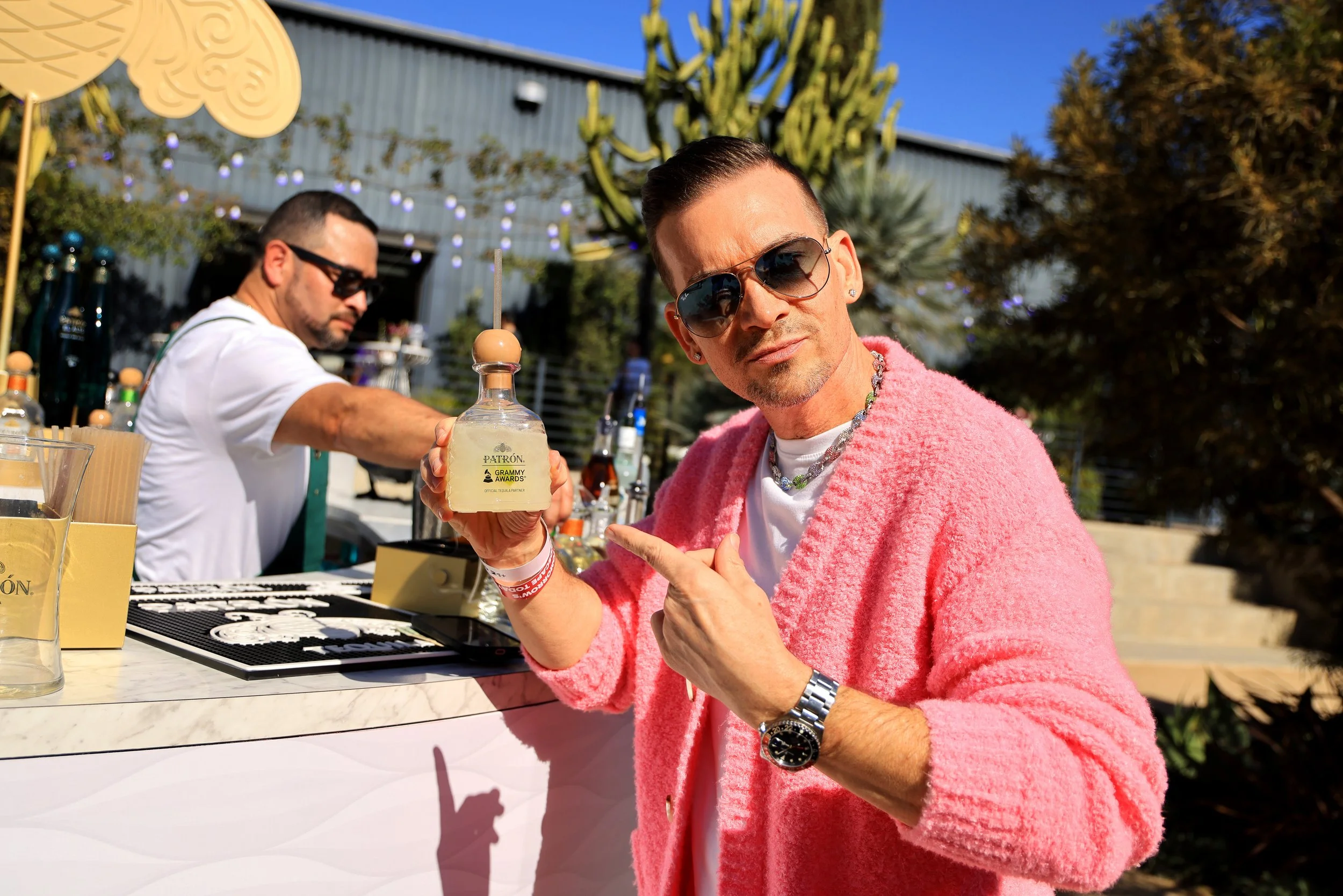 Man wearing pink cardigan, sunglasses, and a silver watch pointing at a bottle of Patrón tequila at an outdoor bar, with another man behind the bar preparing drinks, and outdoor decor including cacti and string lights in the background.