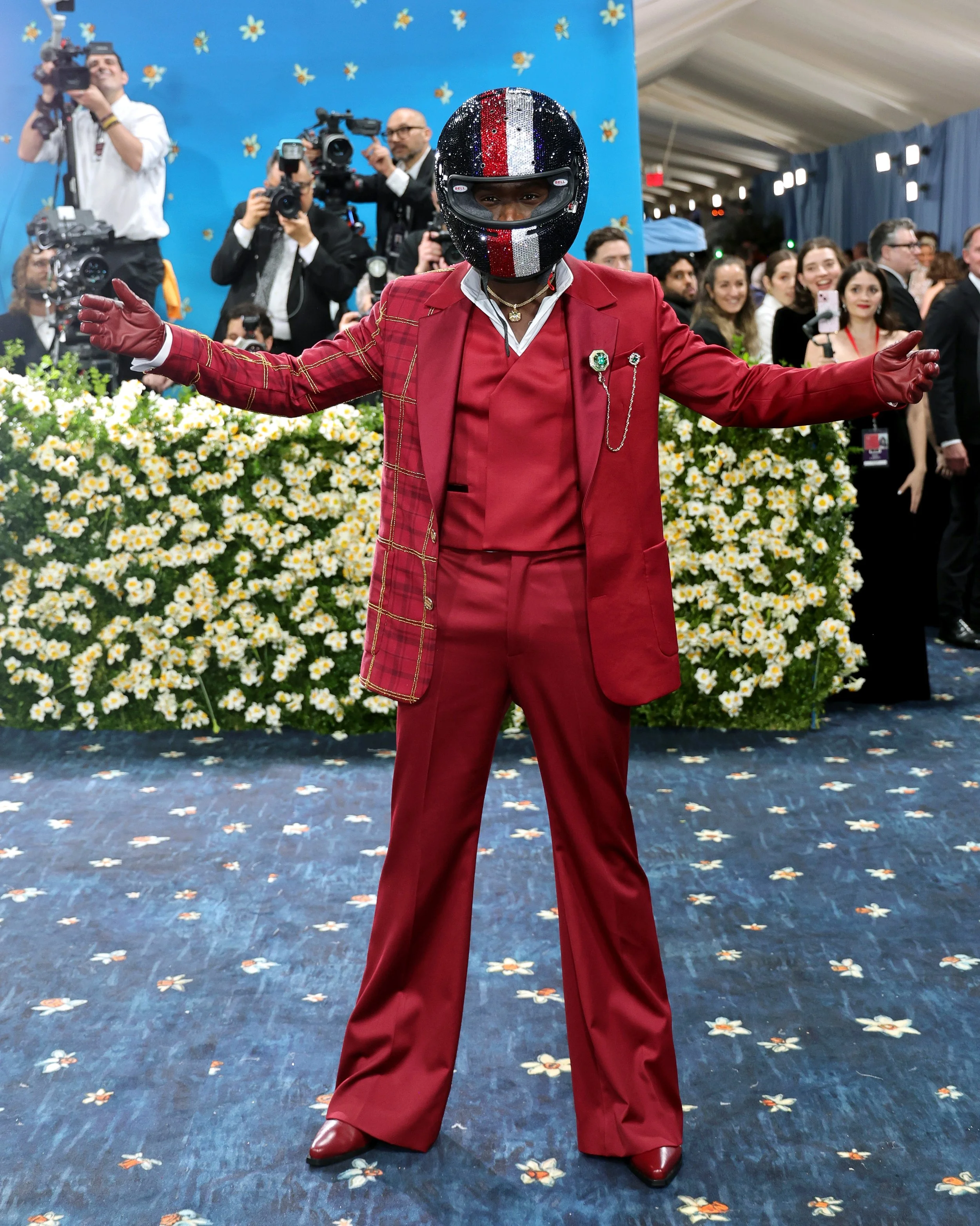 A person dressed in a red suit and gloves, wearing a glittery black helmet with a checkered pattern, stands with arms outstretched at a crowded event with photographers and onlookers, and a backdrop decorated with flowers.
