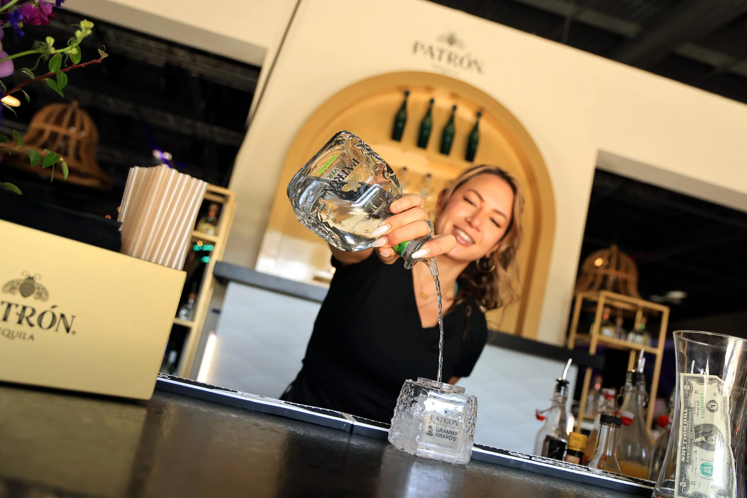 A woman with blonde hair pouring a large glass of water into a glass on a bar counter, with a smile. The bar has bottles, bar tools, and a large glass with a cash bill in it. A box labeled Patrón Tequila and a container with drinking straws are visib