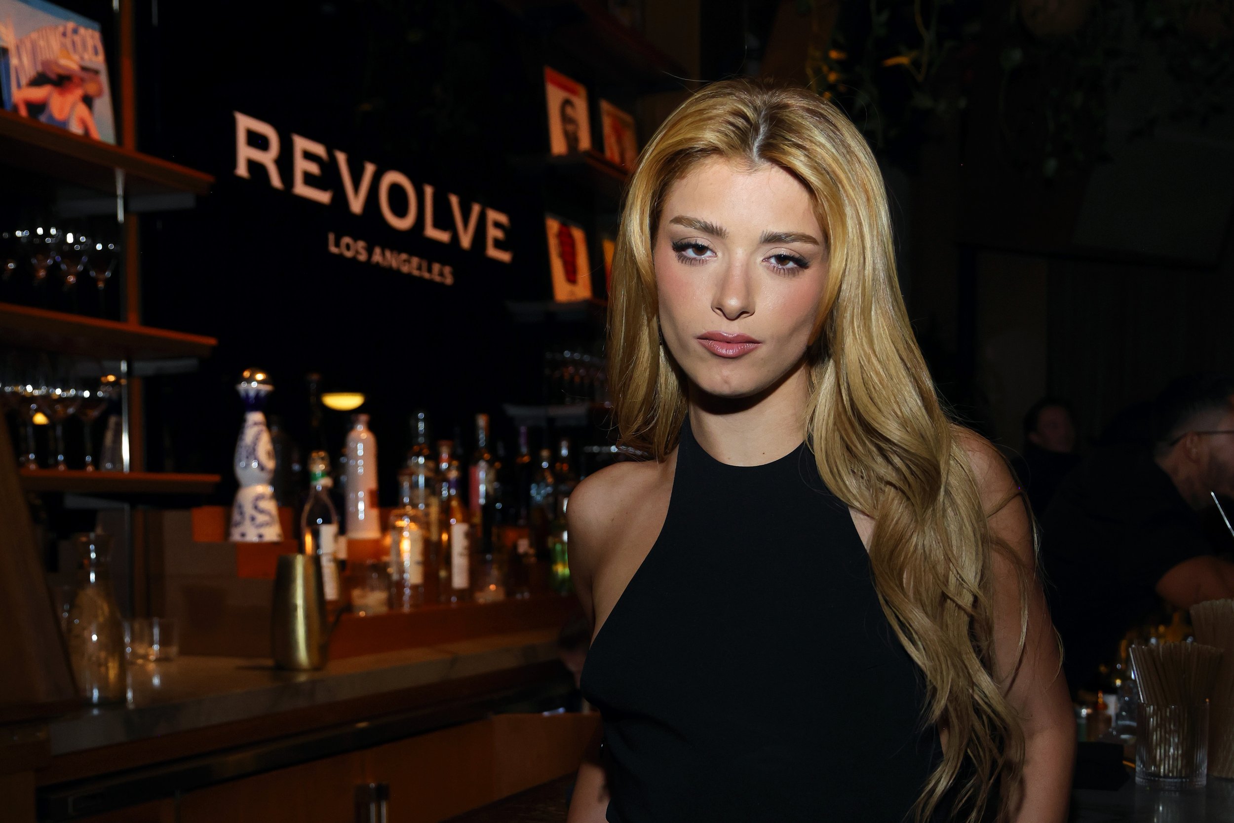 A woman with long wavy blonde hair and light makeup wearing a black sleeveless top stands in a bar with a sign that reads 'REVOLVE LOS ANGELES' behind her.