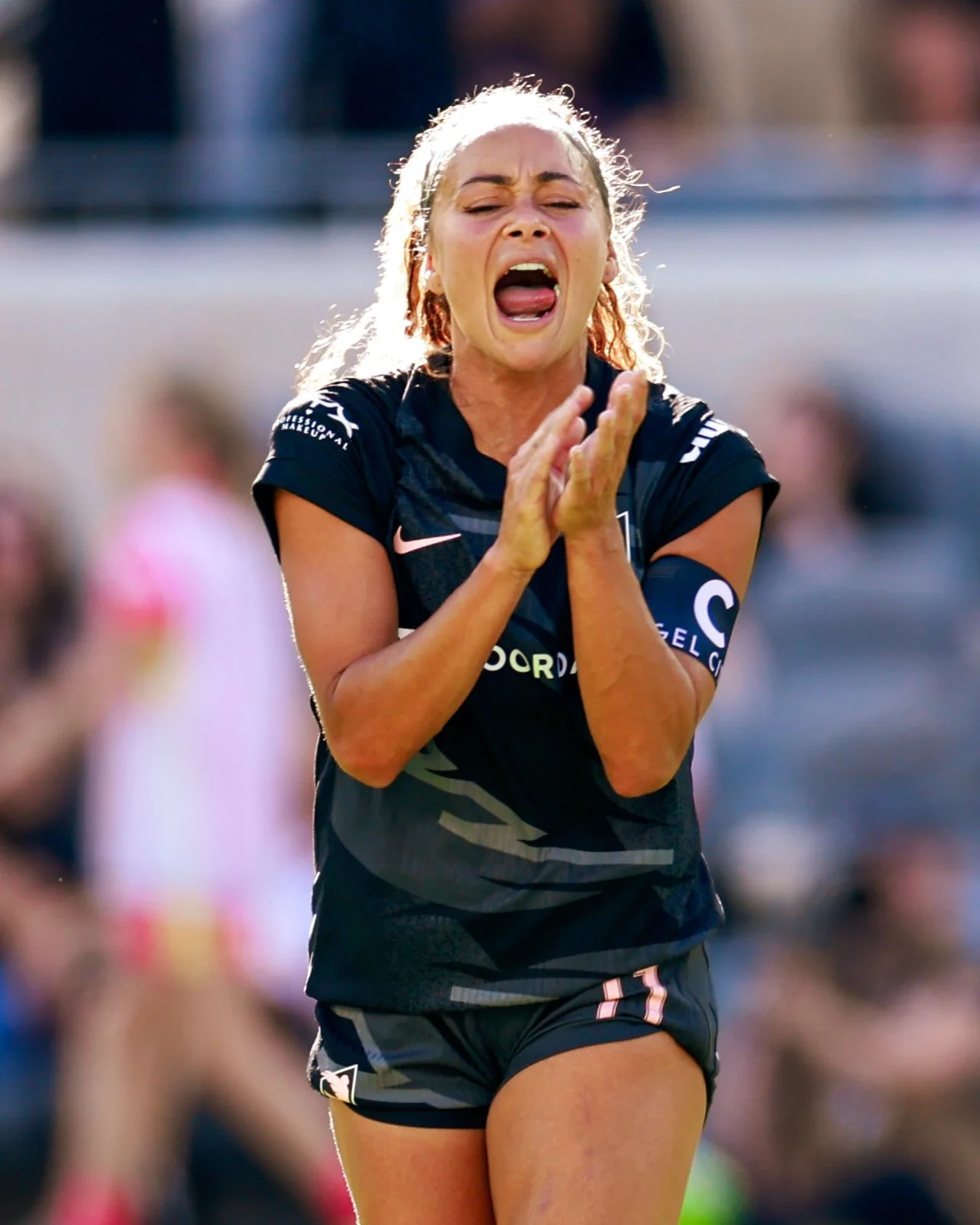 A female athlete wearing a black sports uniform is on a field, appearing emotional with her mouth open and hands together, possibly celebrating or expressing disappointment.