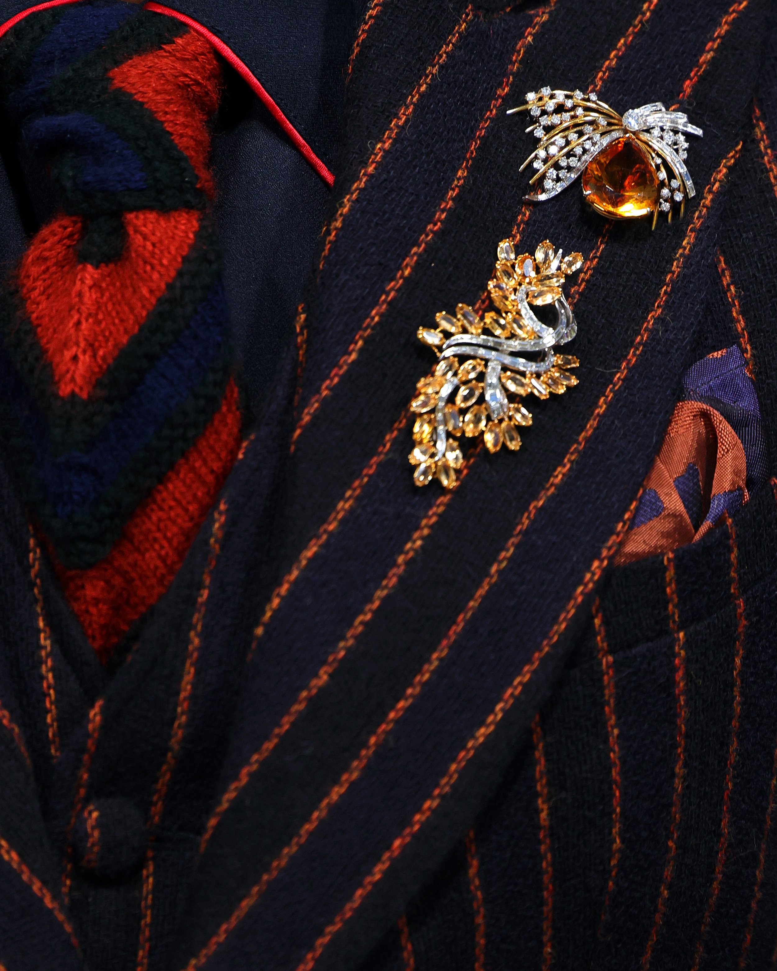 Two ornate brooches, one with a large orange gemstone, the other with a gold and rhinestone floral design, attached to a dark navy pinstripe fabric jacket with a heart-patterned knit sweater underneath.