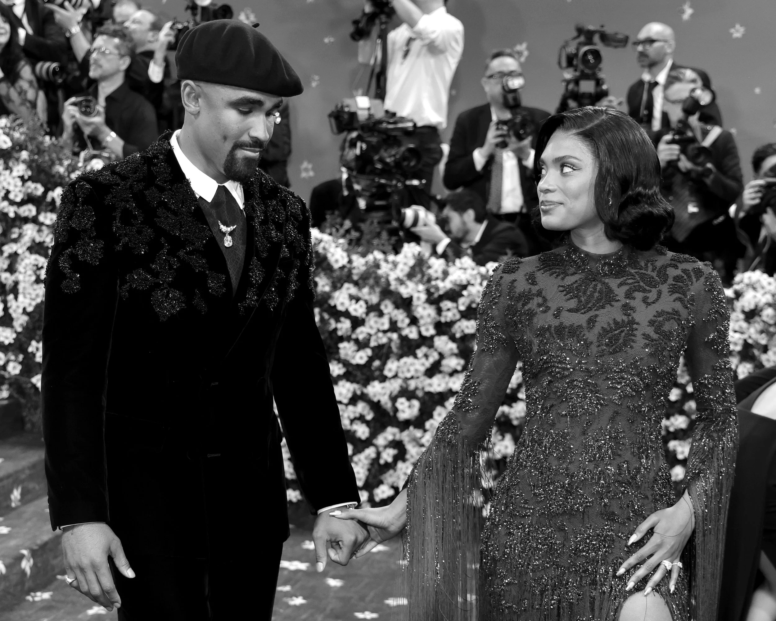 A man and woman holding hands on a red carpet, surrounded by photographers and floral decorations.