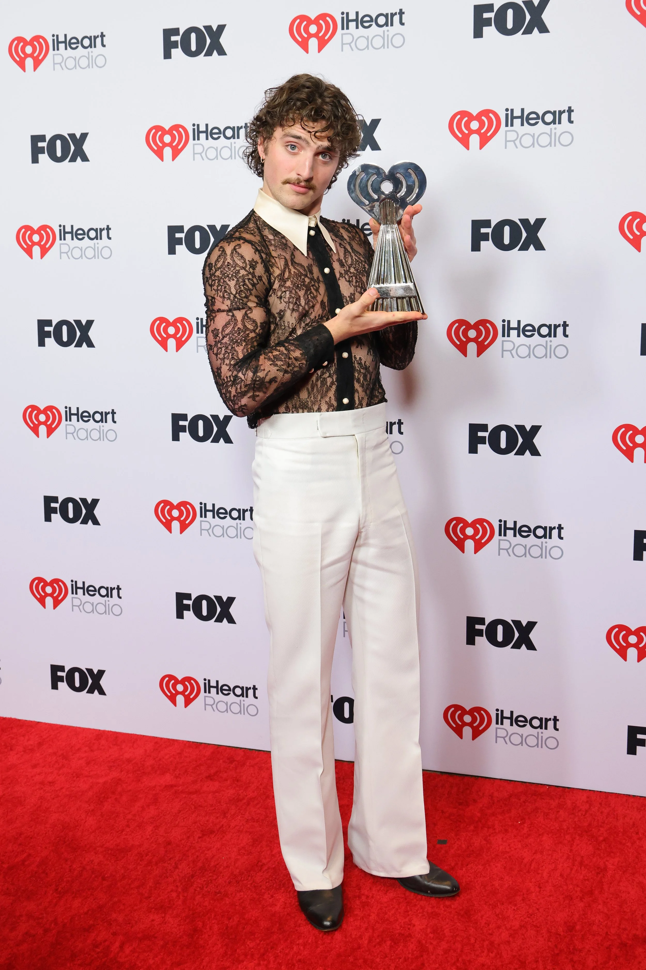A man with curly hair and a mustache holding an award in front of a backdrop with iHeart Radio, FOX logos. He is wearing a black lace shirt, white high-waisted pants, and black shoes.