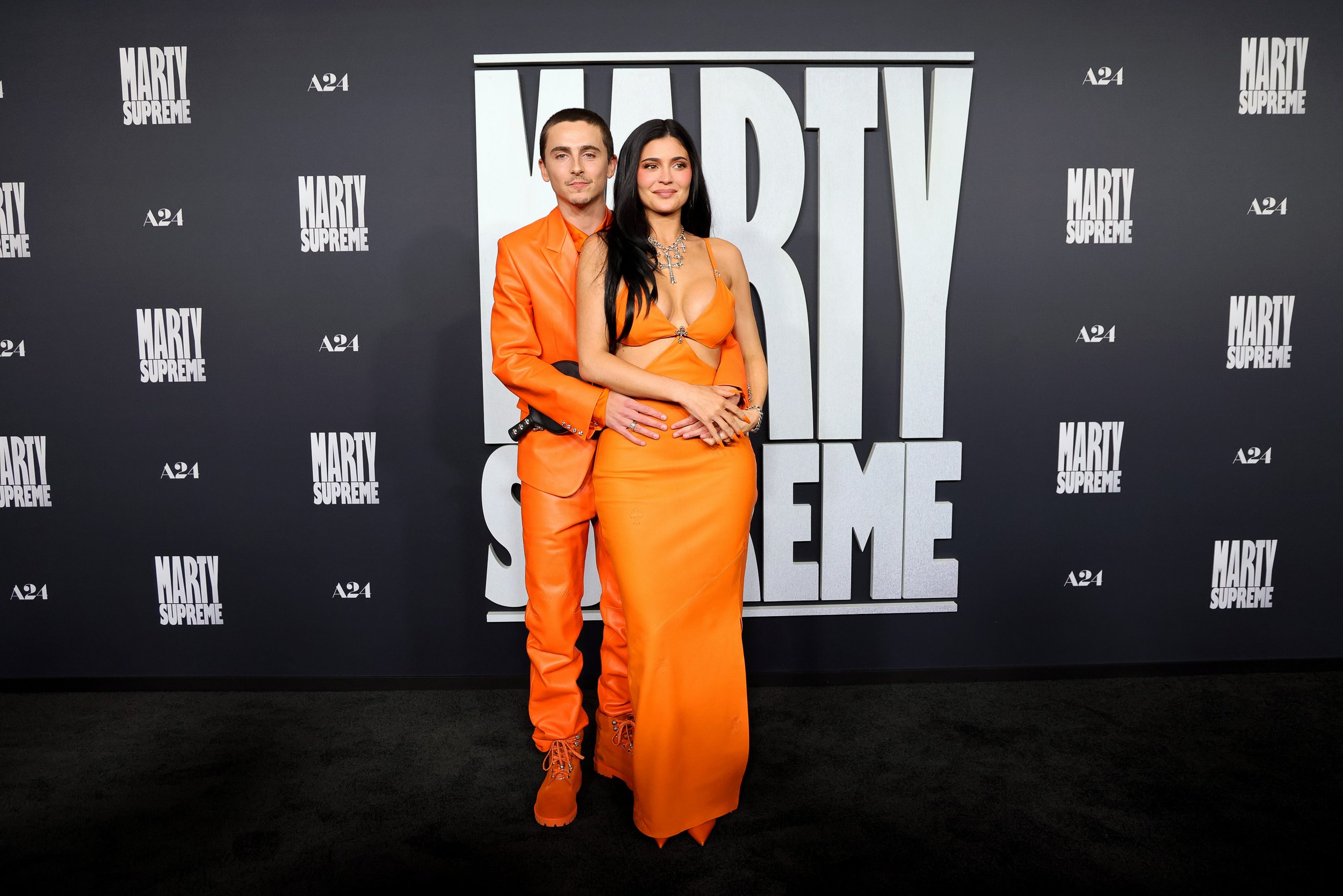 Two people posing together in front of a black backdrop with white text. The man is wearing an orange suit and boots, and the woman is wearing an orange dress with revealing top, jewelry, and long black hair.