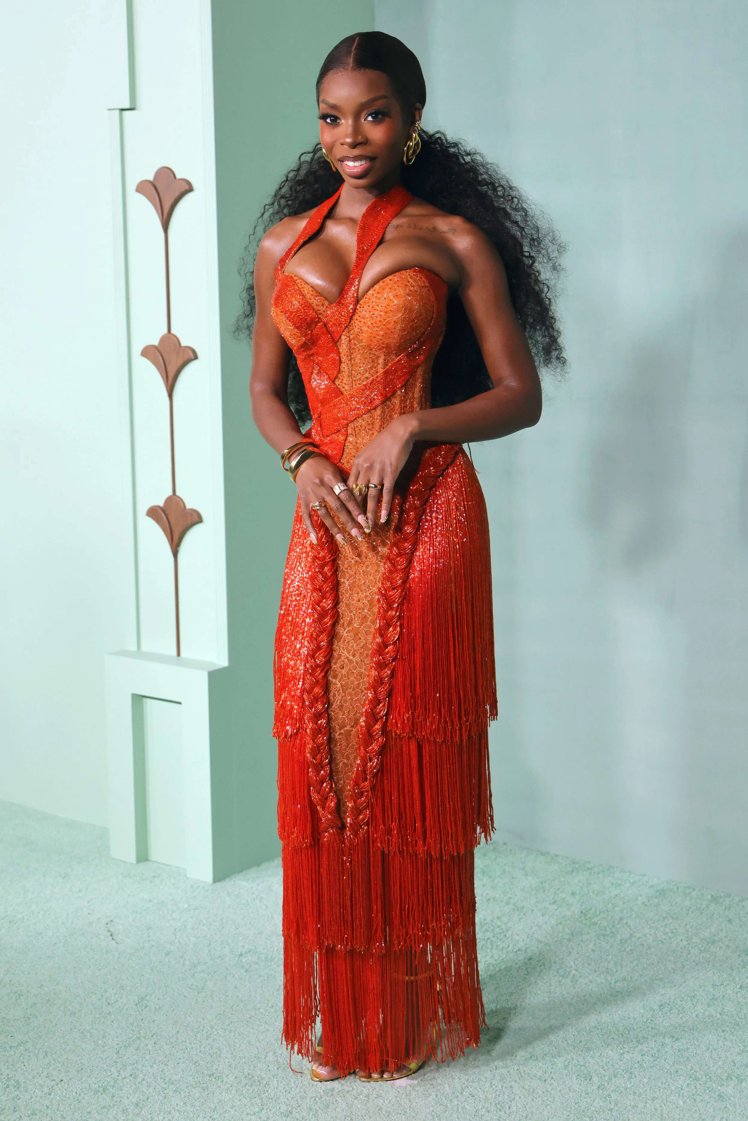 A woman with long, curly black hair wearing a red, shiny, fringe dress with intricate details, standing against a light green wall.