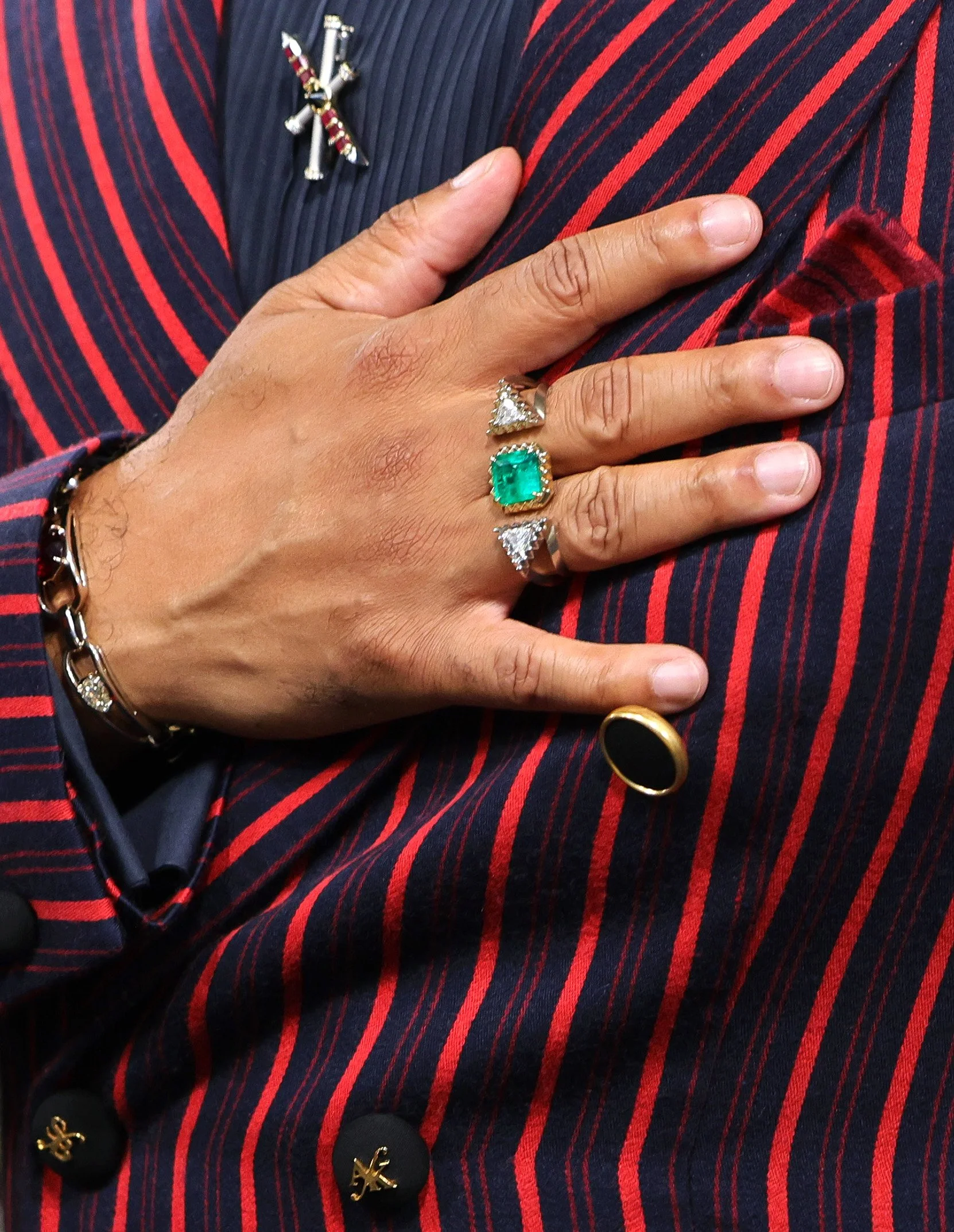 A person's hand with multiple rings rests on their chest, wearing a pinstripe suit with a red and navy design.