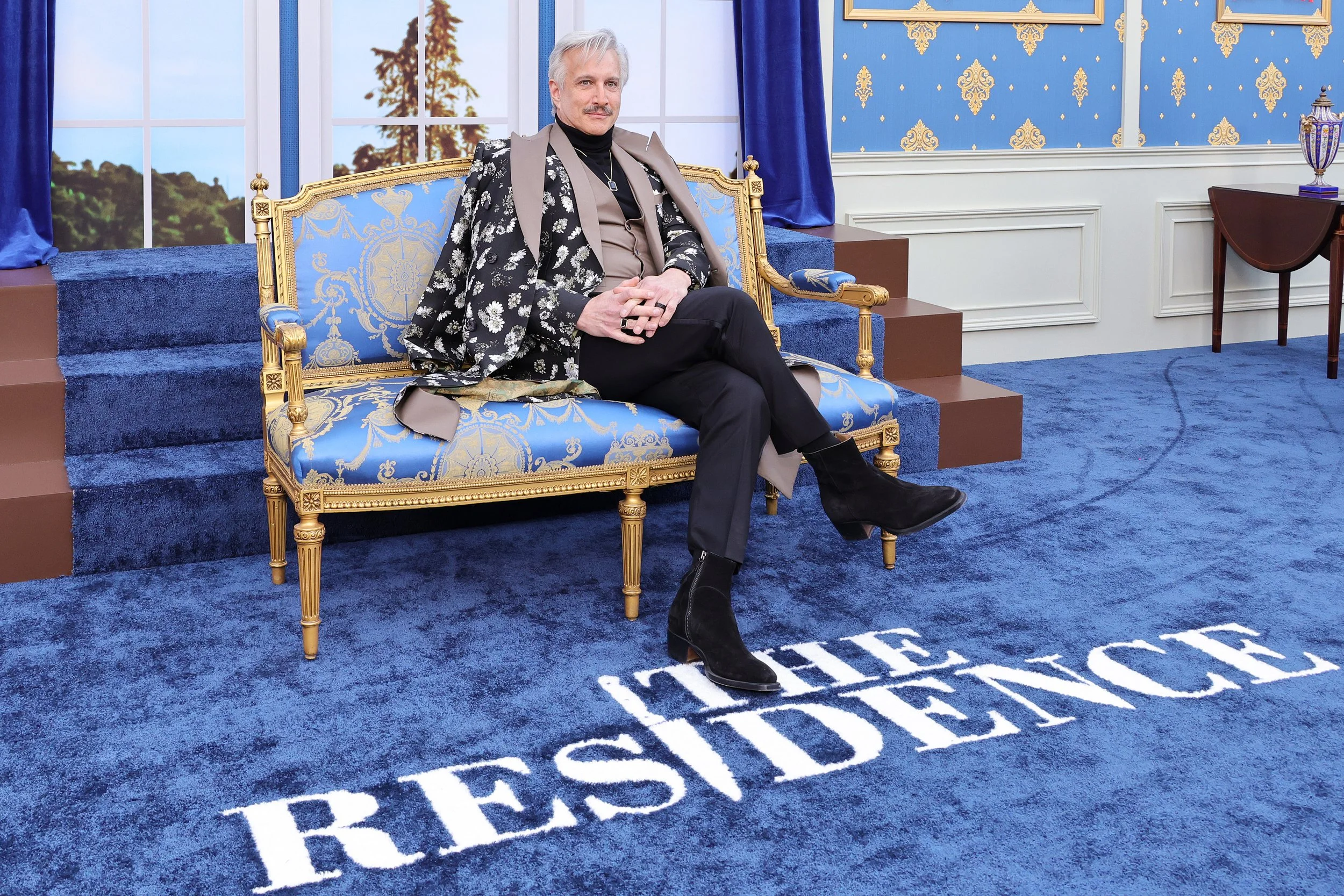 Man sitting on a blue and gold ornate sofa in a decorated room with blue patterned wallpaper and a large window in the background, on a blue carpet with the words "LIFE RESIDENCE" written in white.
