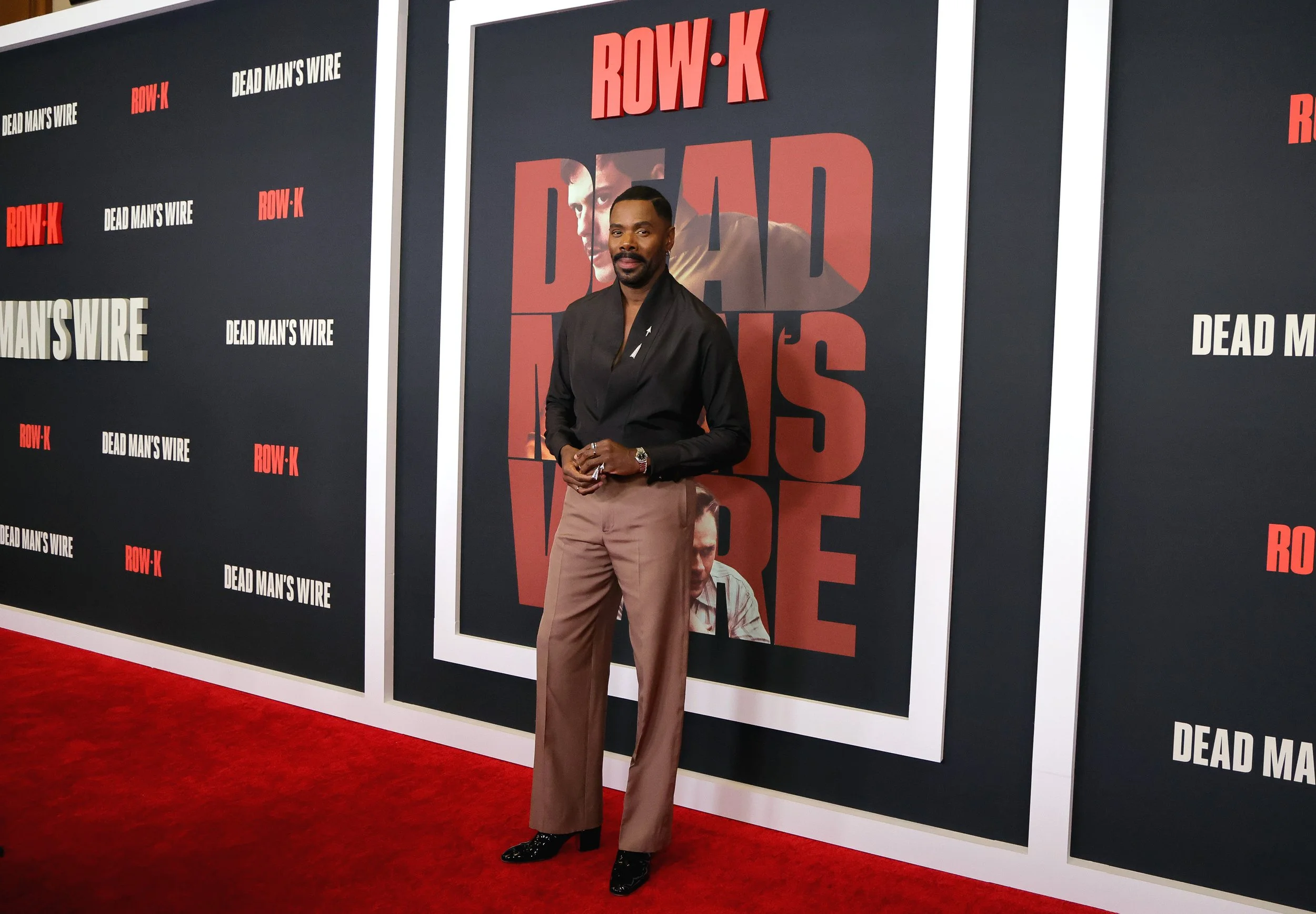 A man posing on a red carpet in front of a promotional backdrop for 'Dead Man's Wire' and 'Row K'. He is wearing a black jacket, beige pants, and black shoes, standing with hands clasped in front of him.