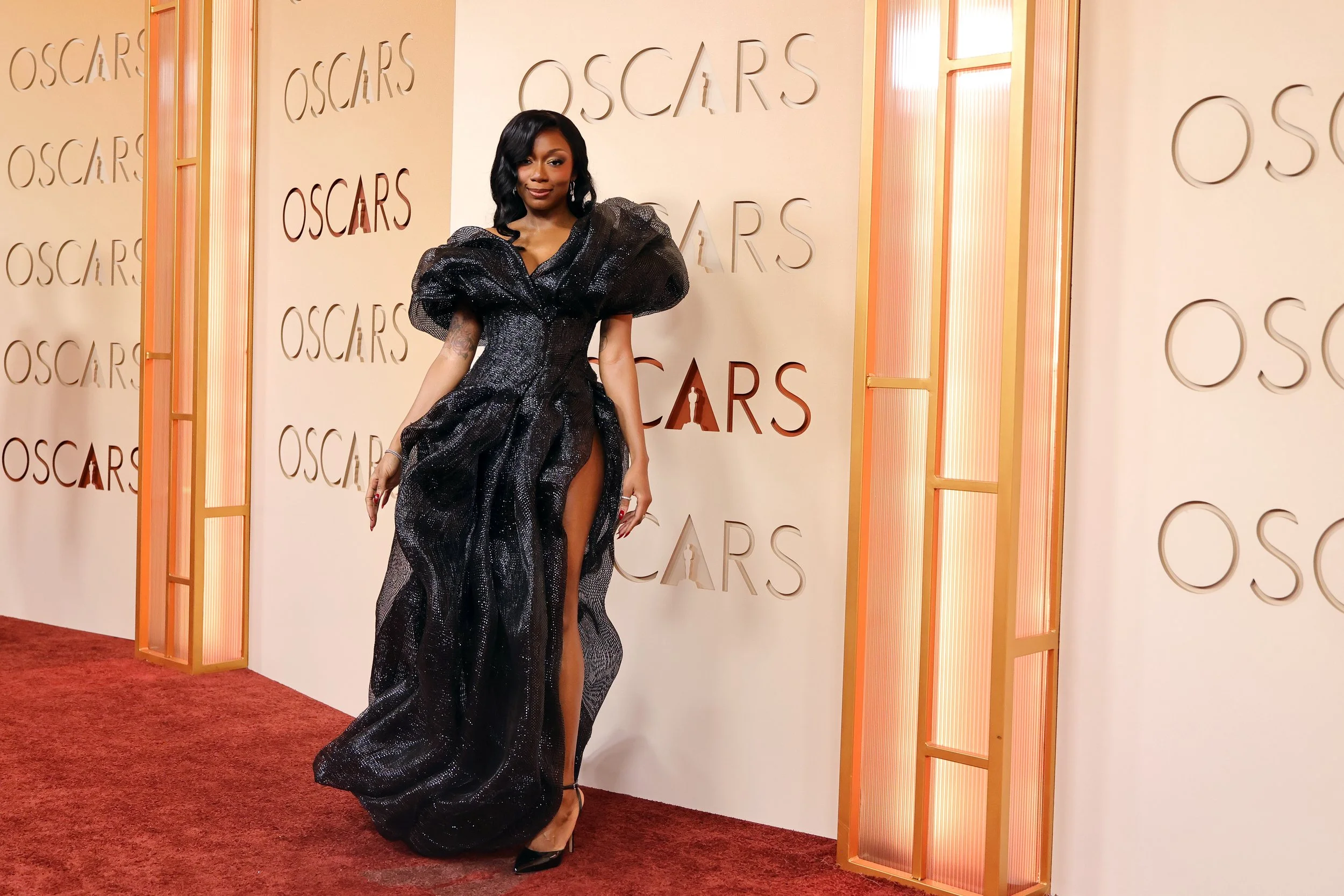 A woman in a black, sparkling gown with a high slit posing on the red carpet at an awards event with 'Oscars' repeated on the backdrop behind her.