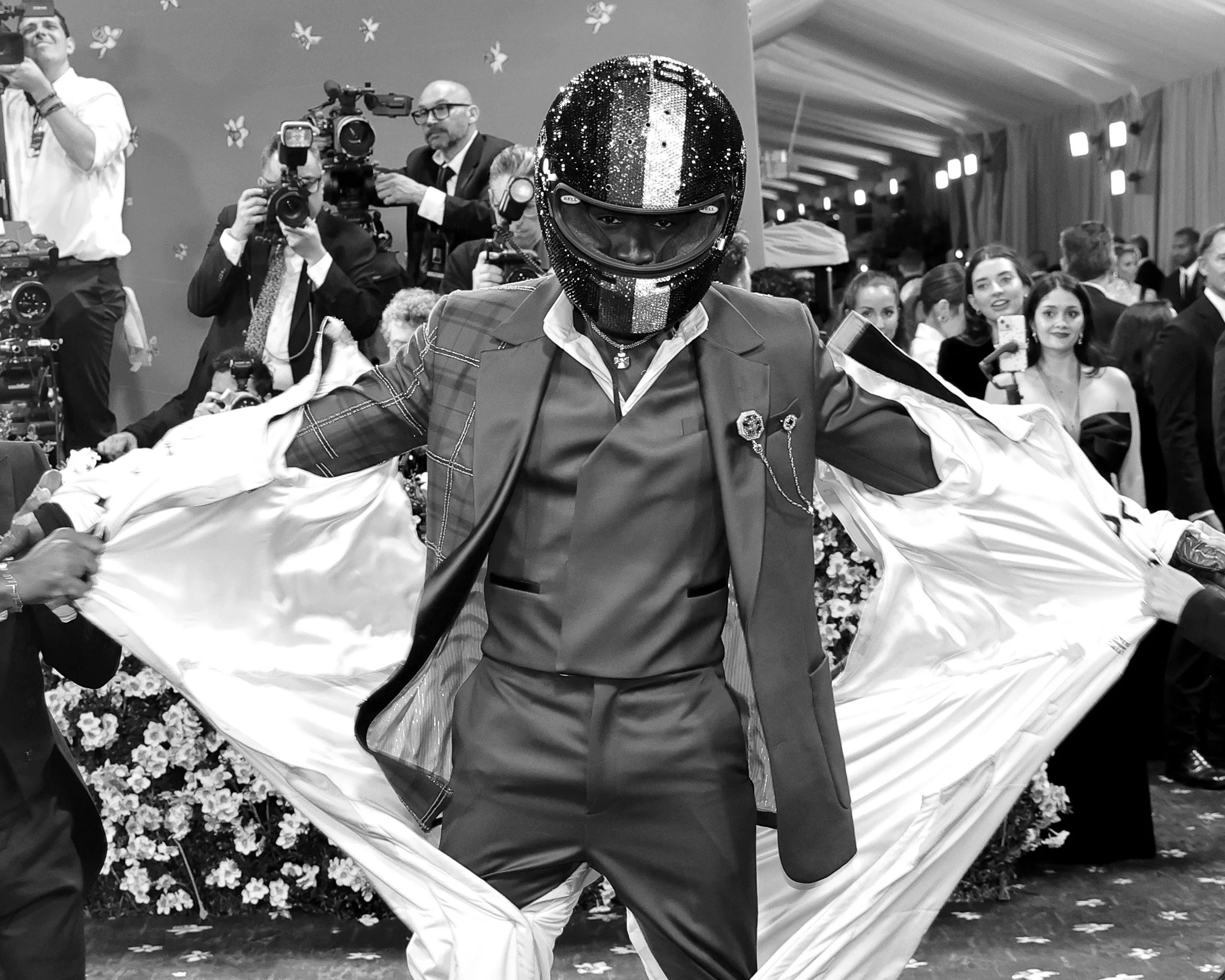 A person wearing a helmet with a shiny, reflective surface, a blazer with decorative pins, and an outfit with contrasting dark and light colors, is walking in a crowded event with photographers and people taking pictures. The person is holding up a s