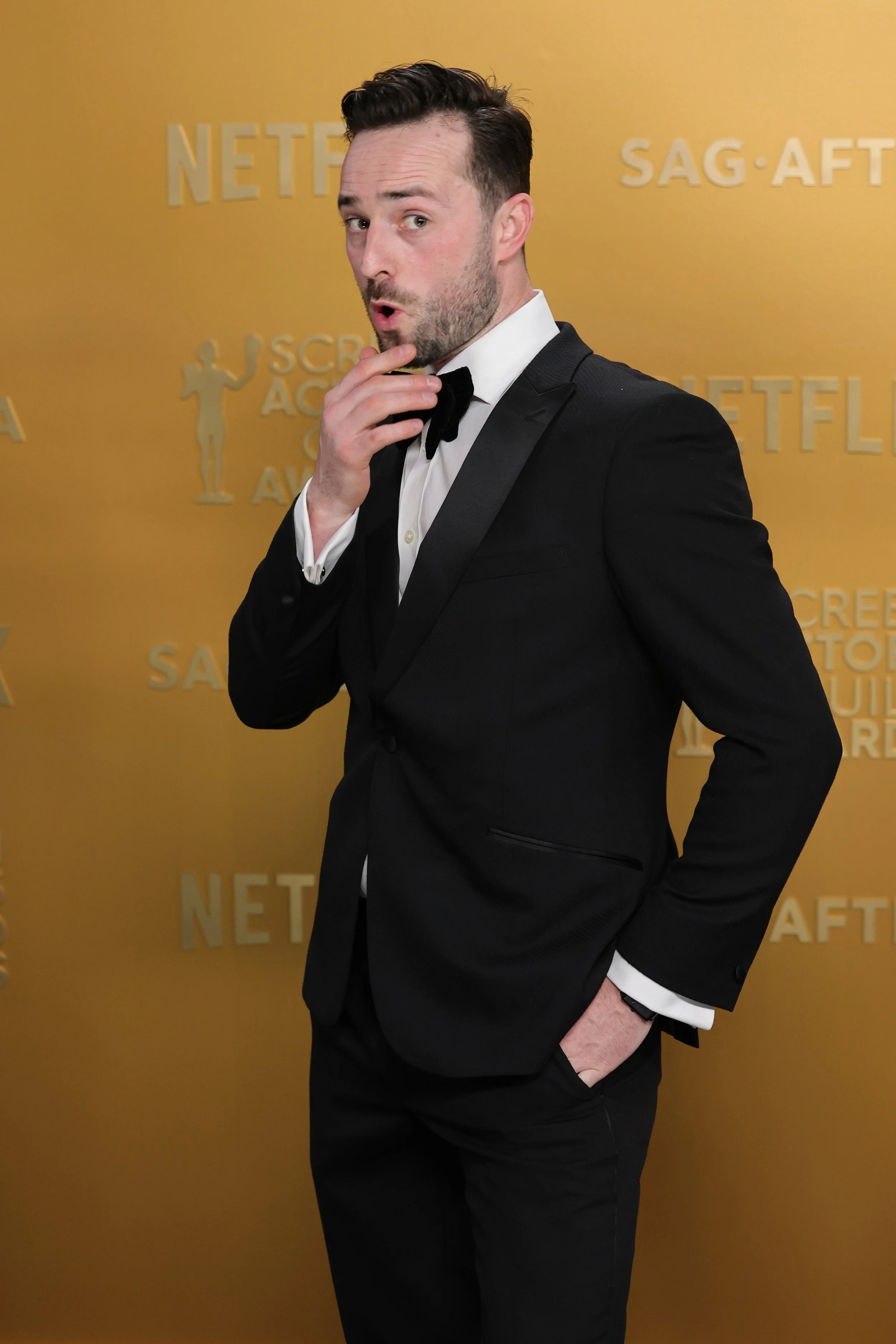 A man in a black tuxedo with a bow tie, standing against a yellow backdrop with Netflix and SAG-AFTRA logos, striking a thoughtful pose with his hand near his chin.