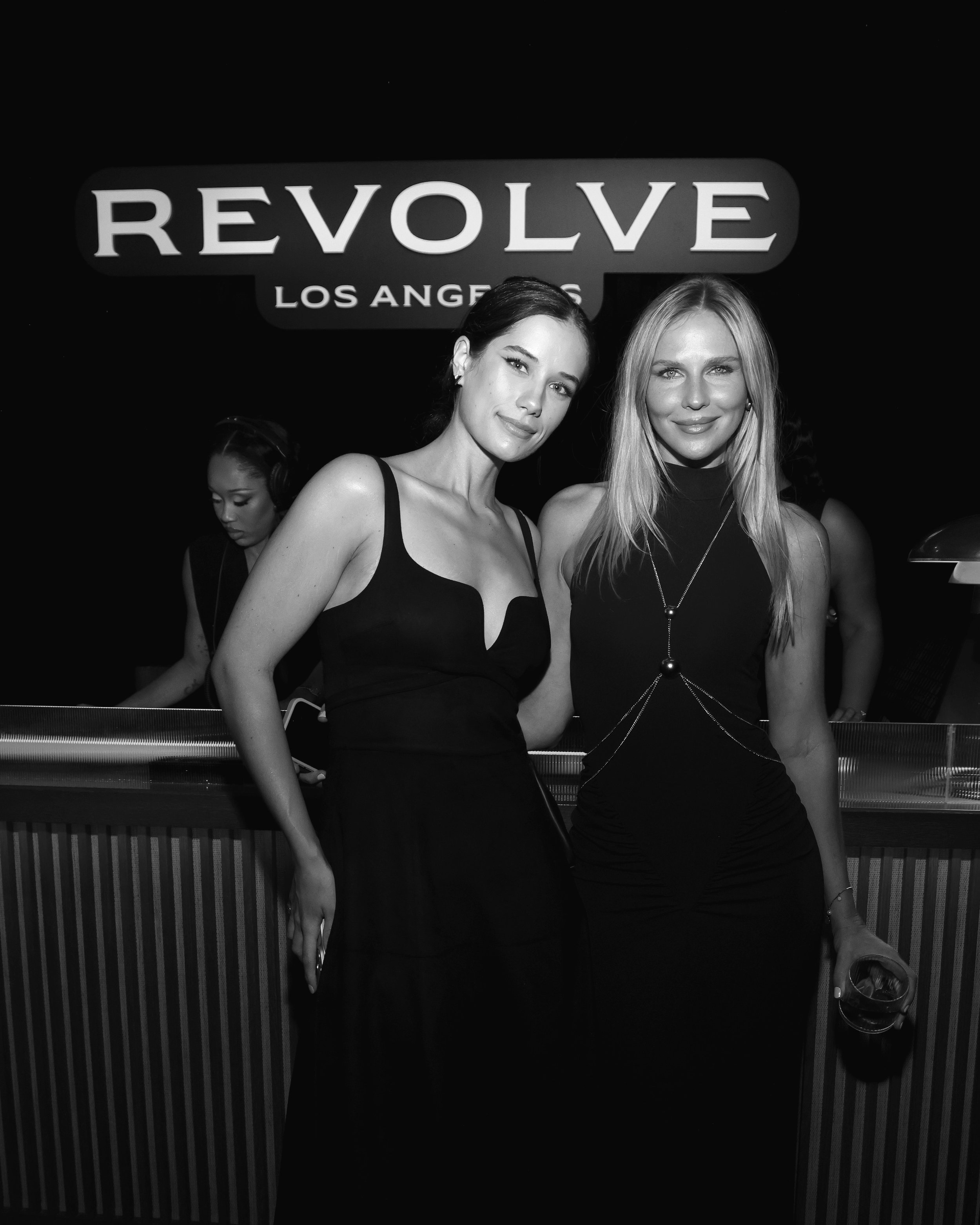 Two women in black dresses at a nightclub, standing in front of a sign that says 'REVOLVE LOS ANGELES'.