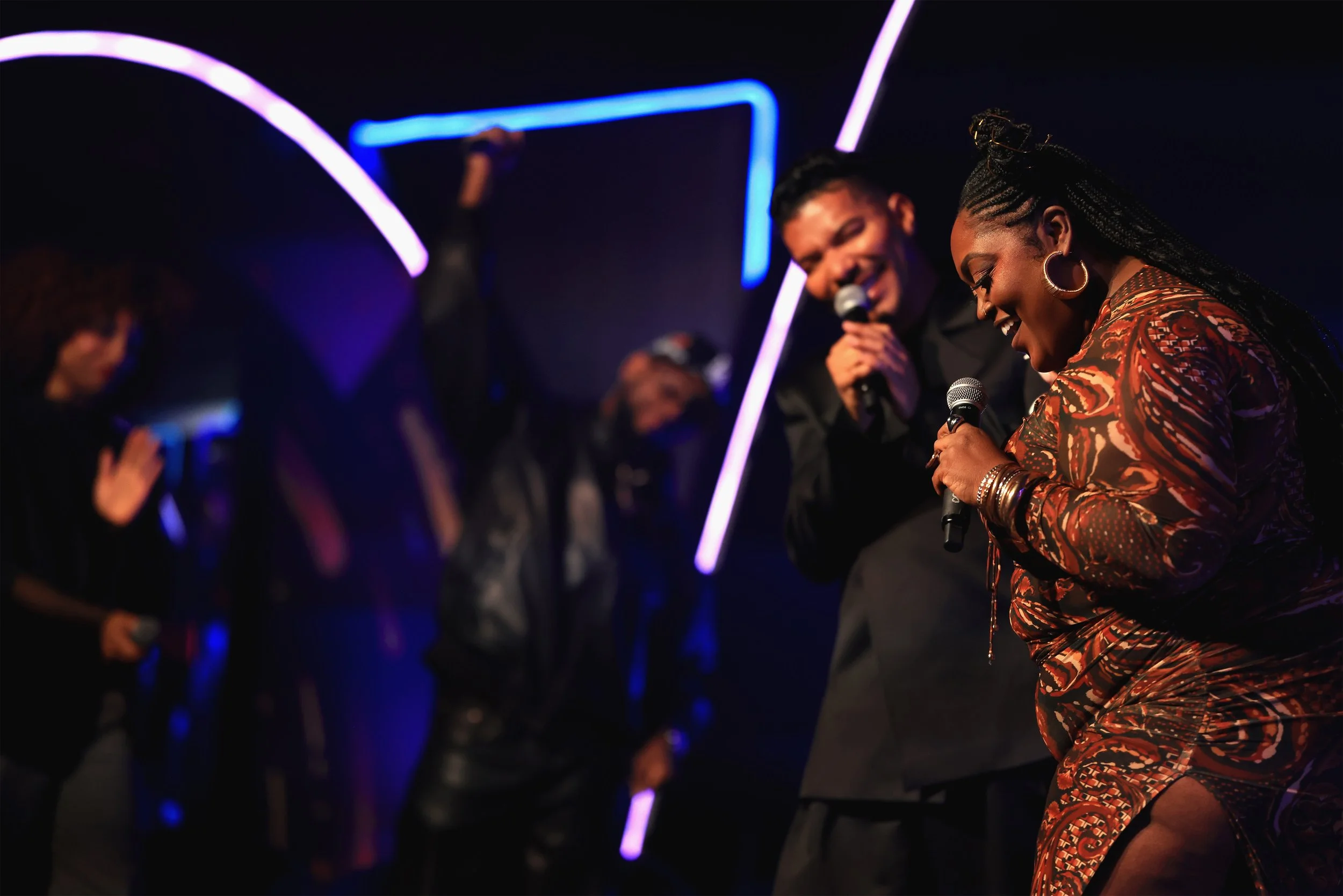 Two women and three men singing into microphones on a stage with neon light decorations in the background.