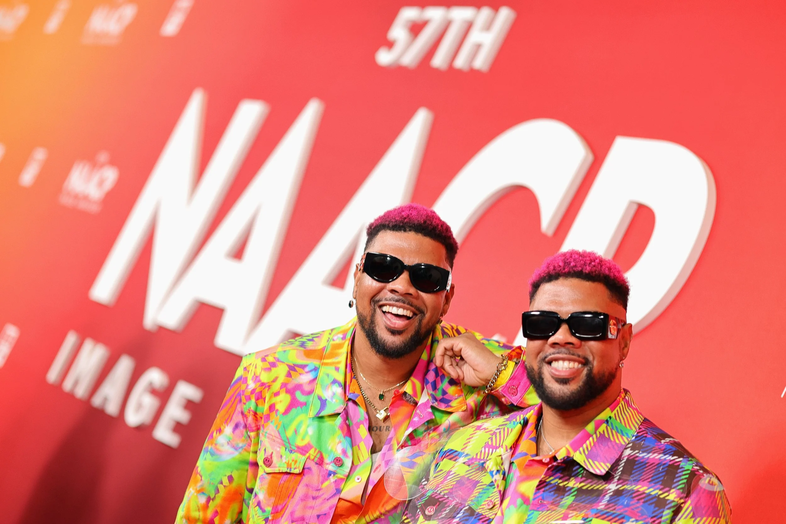 Two men with pink hair wearing colorful patterned shirts and sunglasses, smiling in front of a red background with large white text that says "57th NAACP IMAGE".