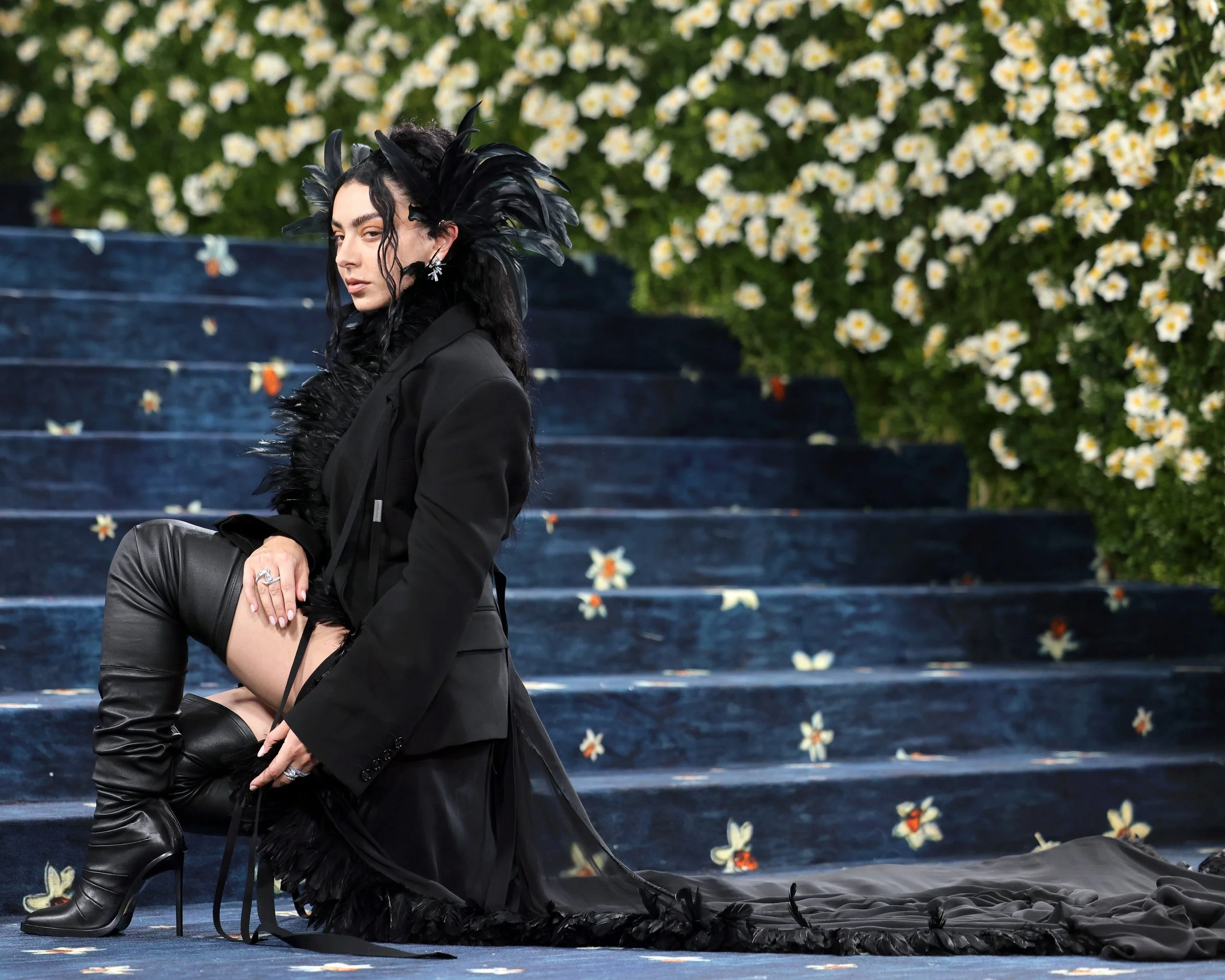 A woman in a black outfit with high thigh boots, sitting on stairs covered with fallen flower petals, with a backdrop of white flowers and greenery.
