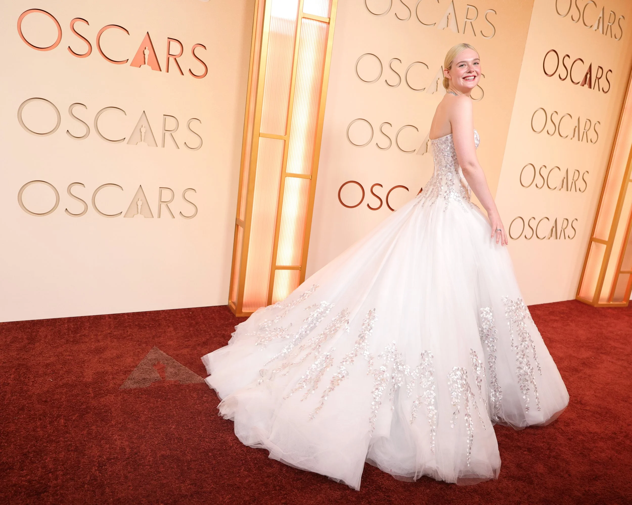 A woman in a white, strapless, ball gown with floral embroidery, standing on a red carpet at an Oscars event, smiling and looking over her shoulder.