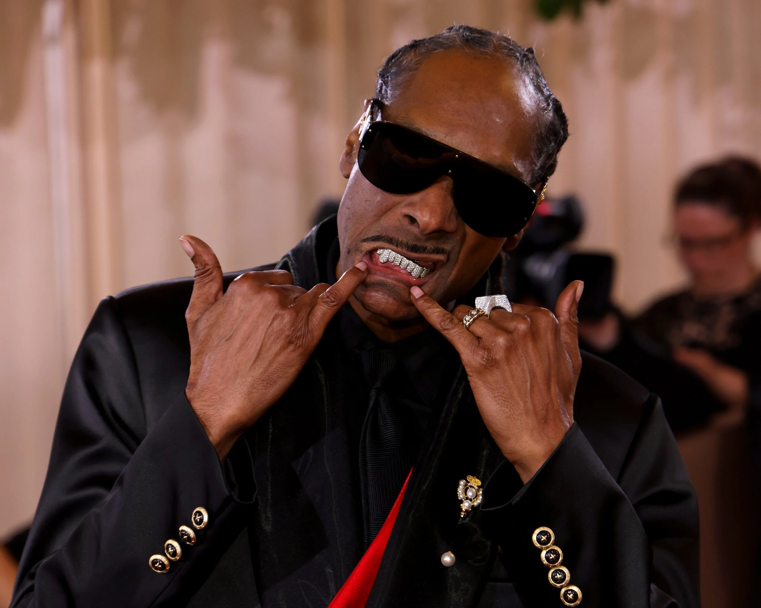 A man wearing dark sunglasses, a black leather jacket with gold buttons, and over-the-top jewelry, showing a grill with diamonds on his teeth. He is making a 'rock on' hand gesture with both hands, with a playful facial expression.