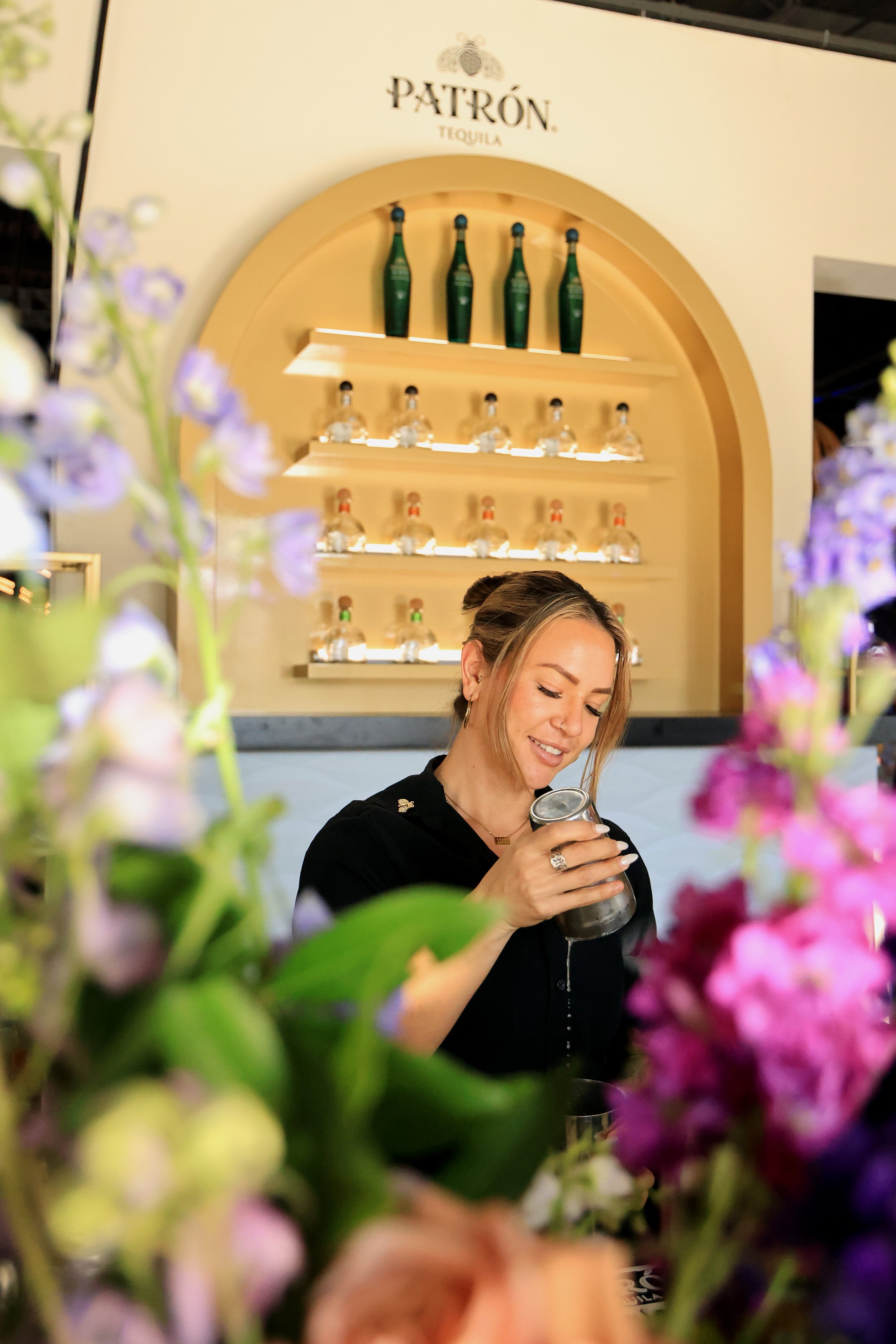 A woman smiling and pouring a drink behind a floral arrangement at a Patrón tequila display.