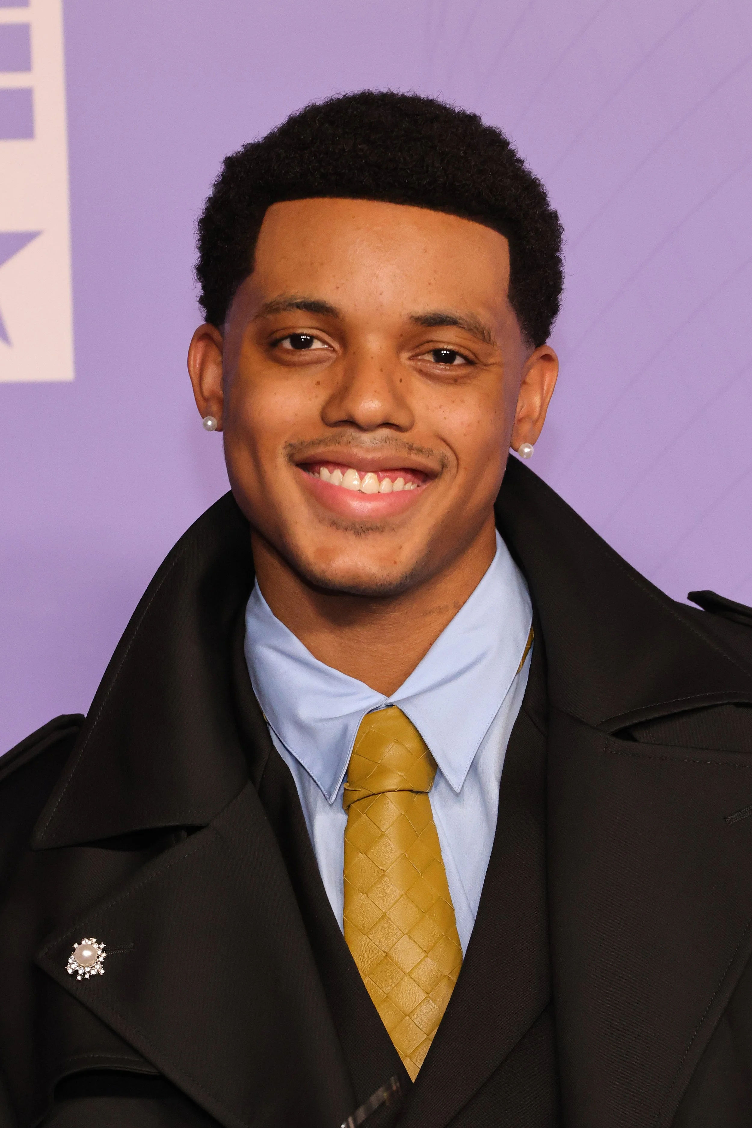 A young man smiling in a professional outfit; he wears a black blazer, light blue shirt, gold tie, and pearl earrings, with a light purple background.