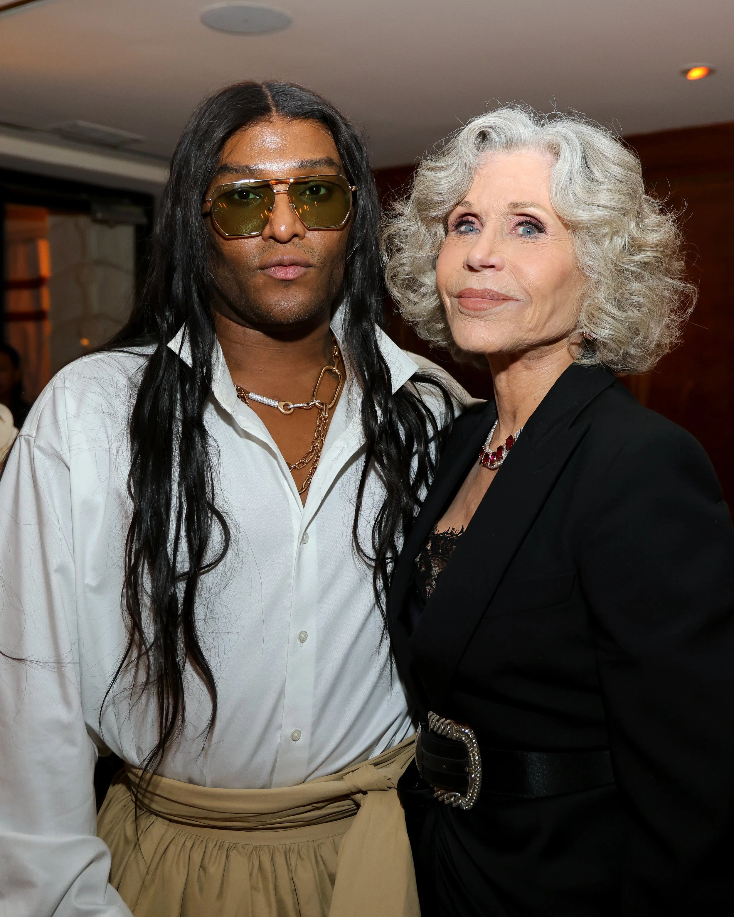 A man with long black hair, wearing sunglasses, a white shirt, and layered necklaces, standing next to an older woman with curly gray hair, wearing a black blazer and a jeweled necklace, at an indoor event.