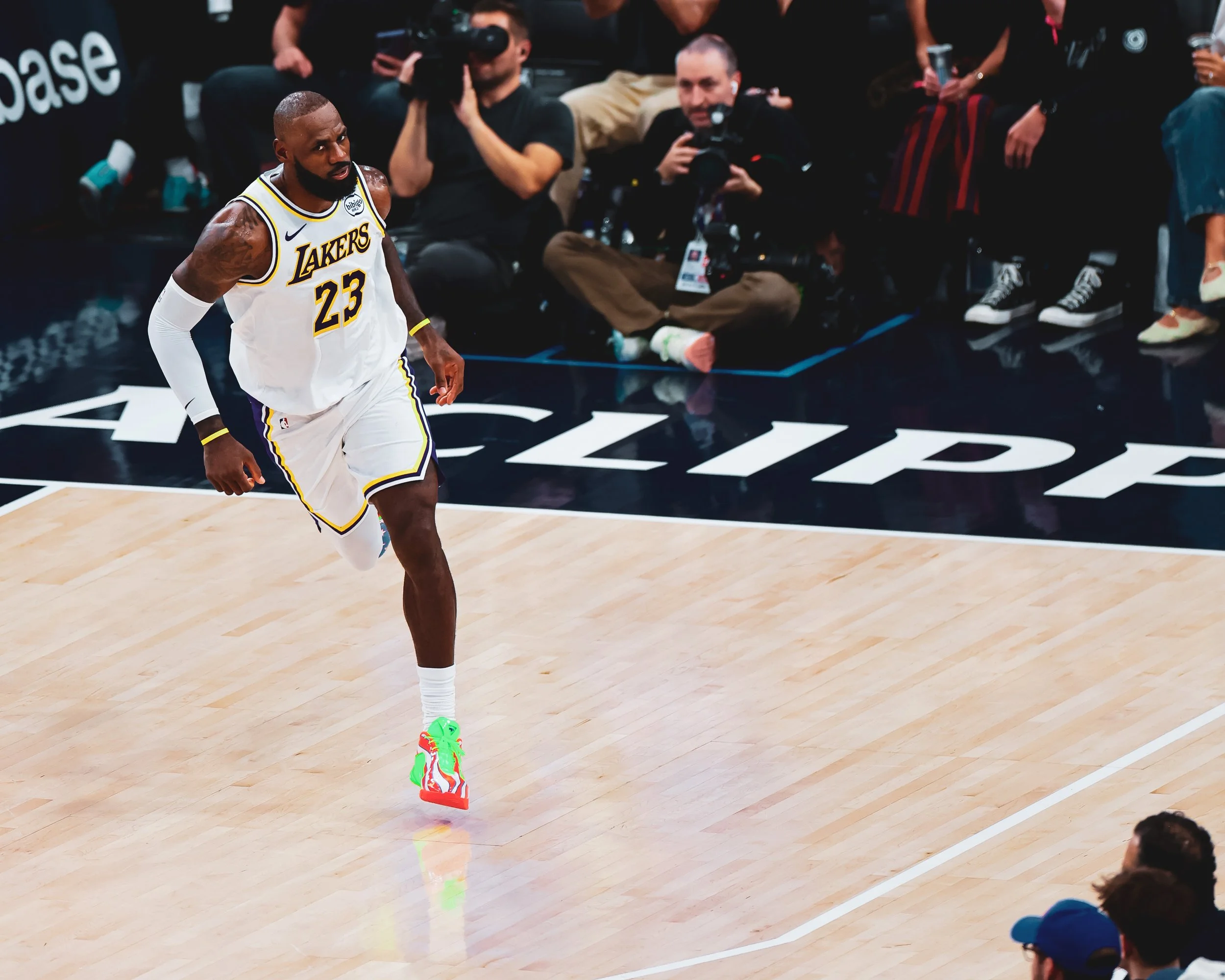 LeBron James, wearing a Los Angeles Lakers uniform with the number 23, jogging on a basketball court during a game. Spectators and photographers sit in the background.