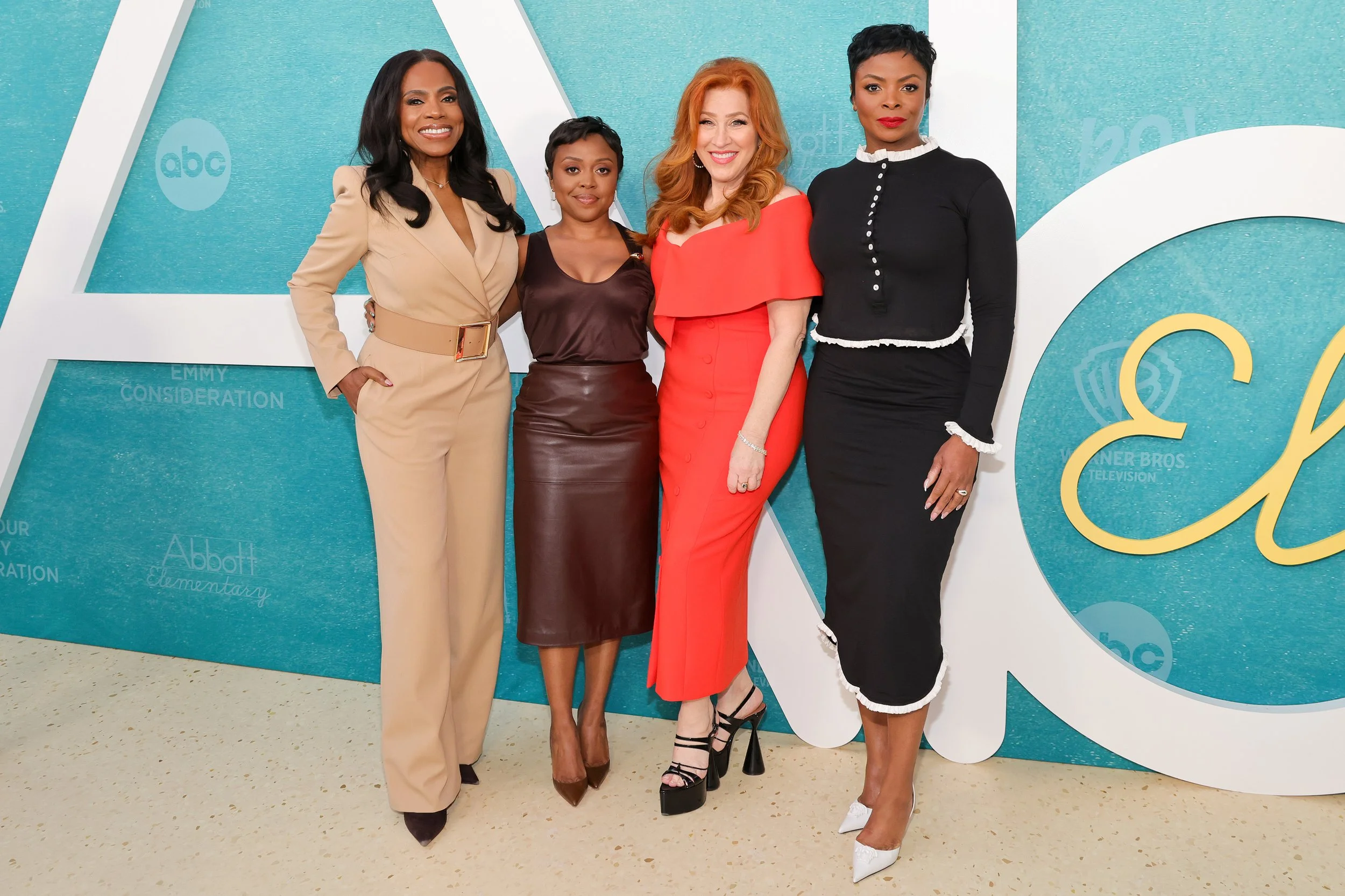 Four women dressed in formal attire standing together outdoors in front of a large teal-colored backdrop with white lettering. They are smiling and posing for a photo.