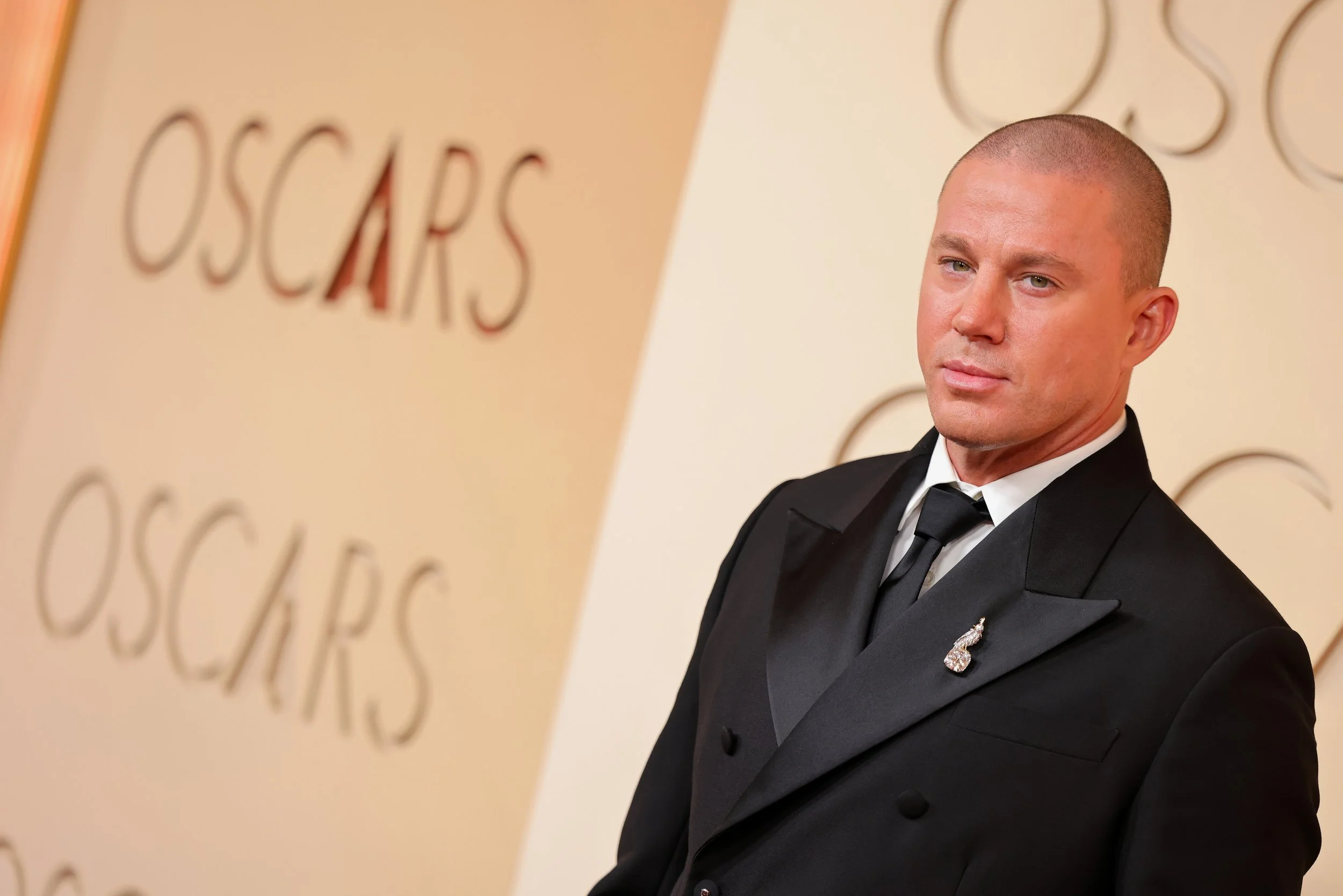 A man in a black tuxedo with a white shirt and black tie standing at an awards event, with a backdrop showing the phrase 'Oscars' repeatedly.