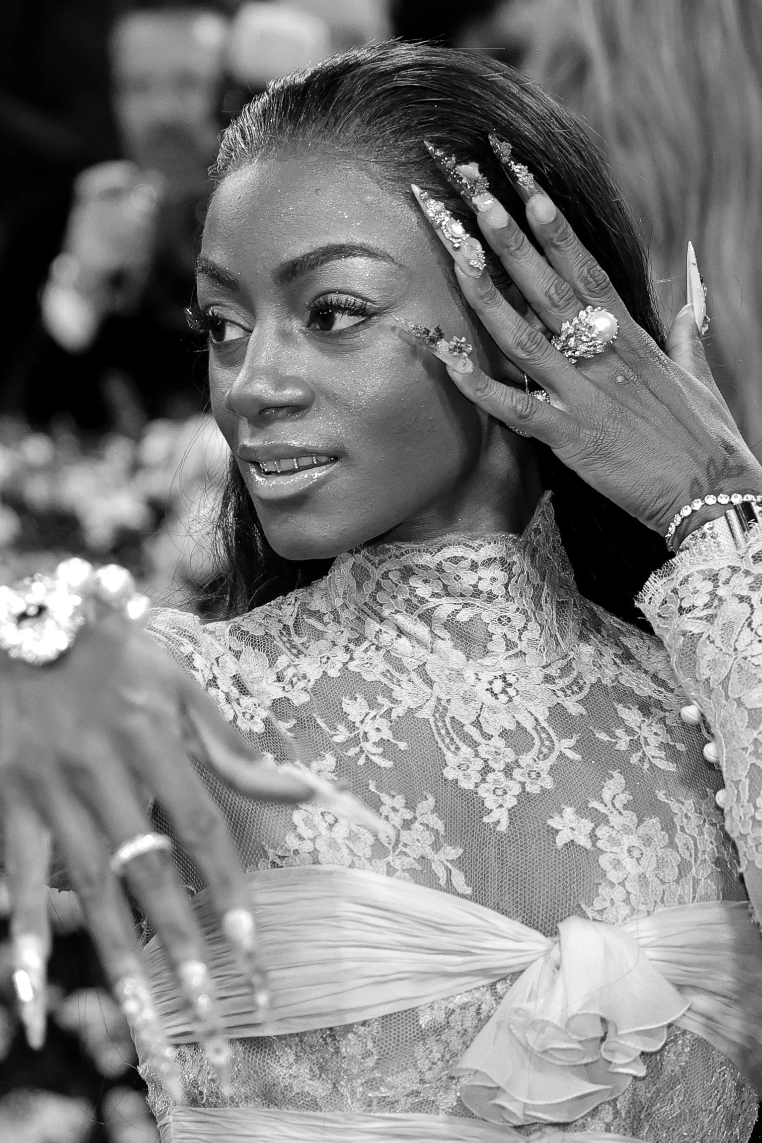 A woman with long dark hair, wearing a lace dress with floral embroidery, is touching her face with her left hand, which is adorned with earrings, rings, and a bracelet. She appears to be at a celebration or special event.