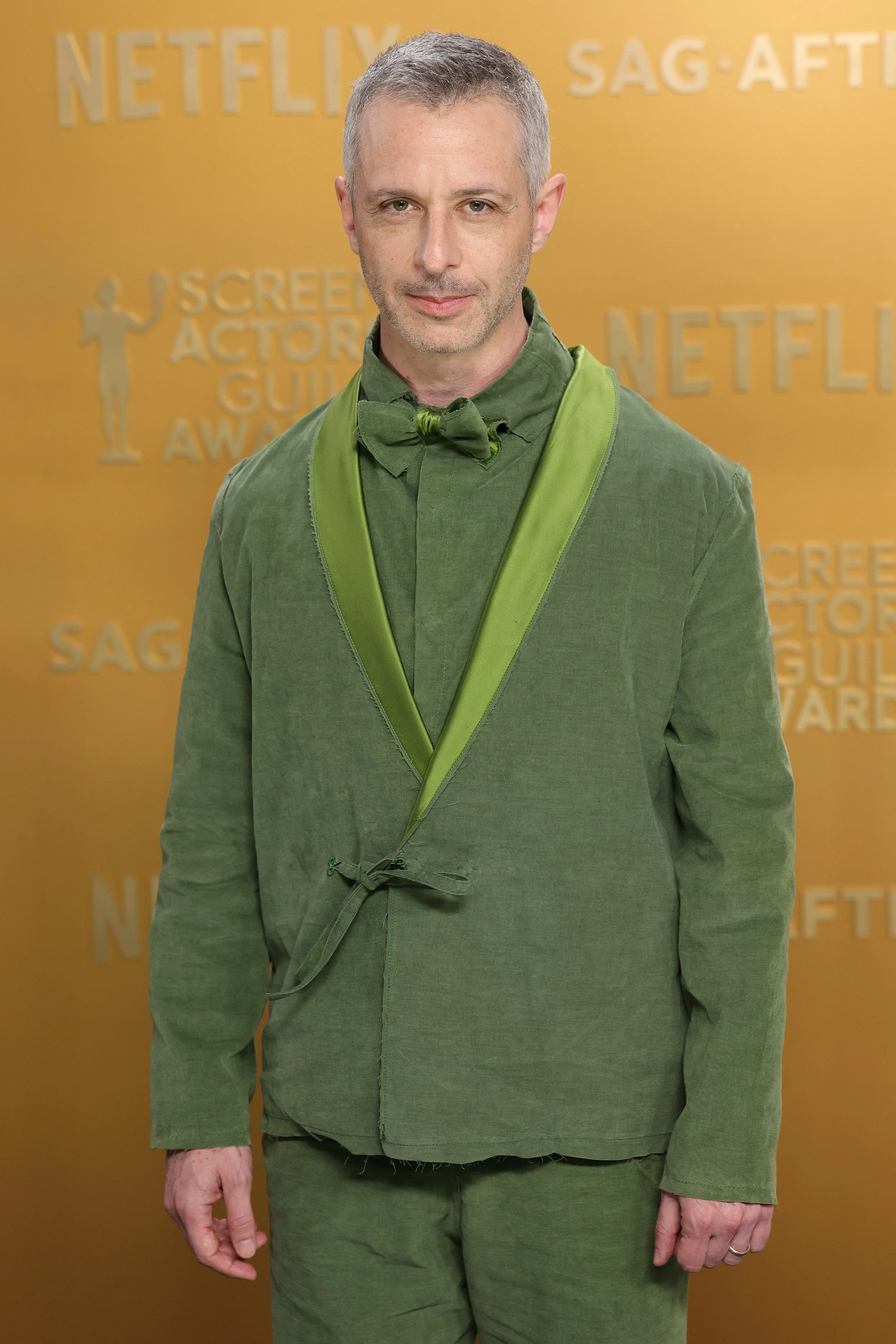 Man with short gray hair and stubble wearing a green suit with a matching green bow tie, standing in front of a yellow background with text, at an awards event.
