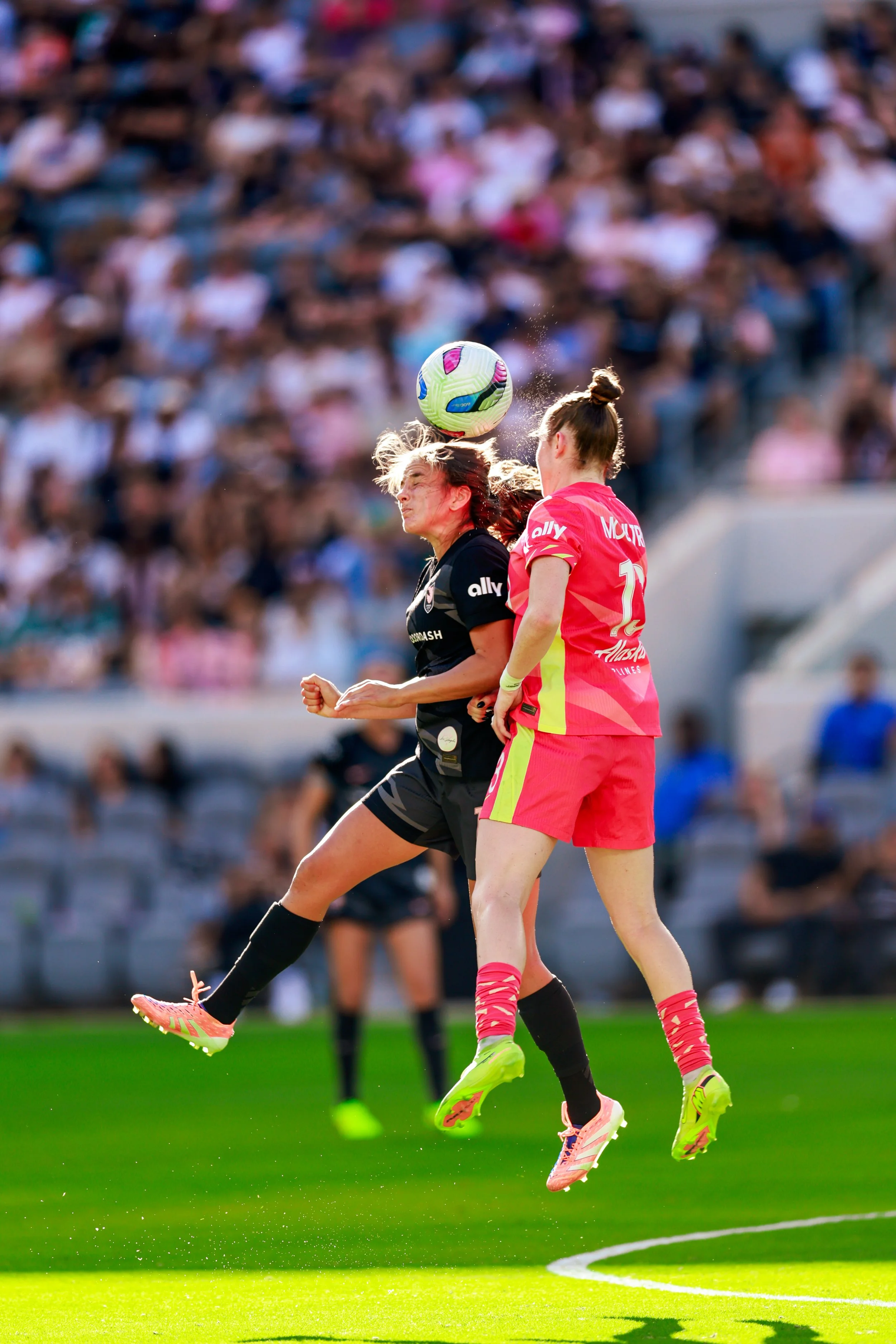 Two female soccer players jumping to head the ball during a match, with a blurred crowd in the background.