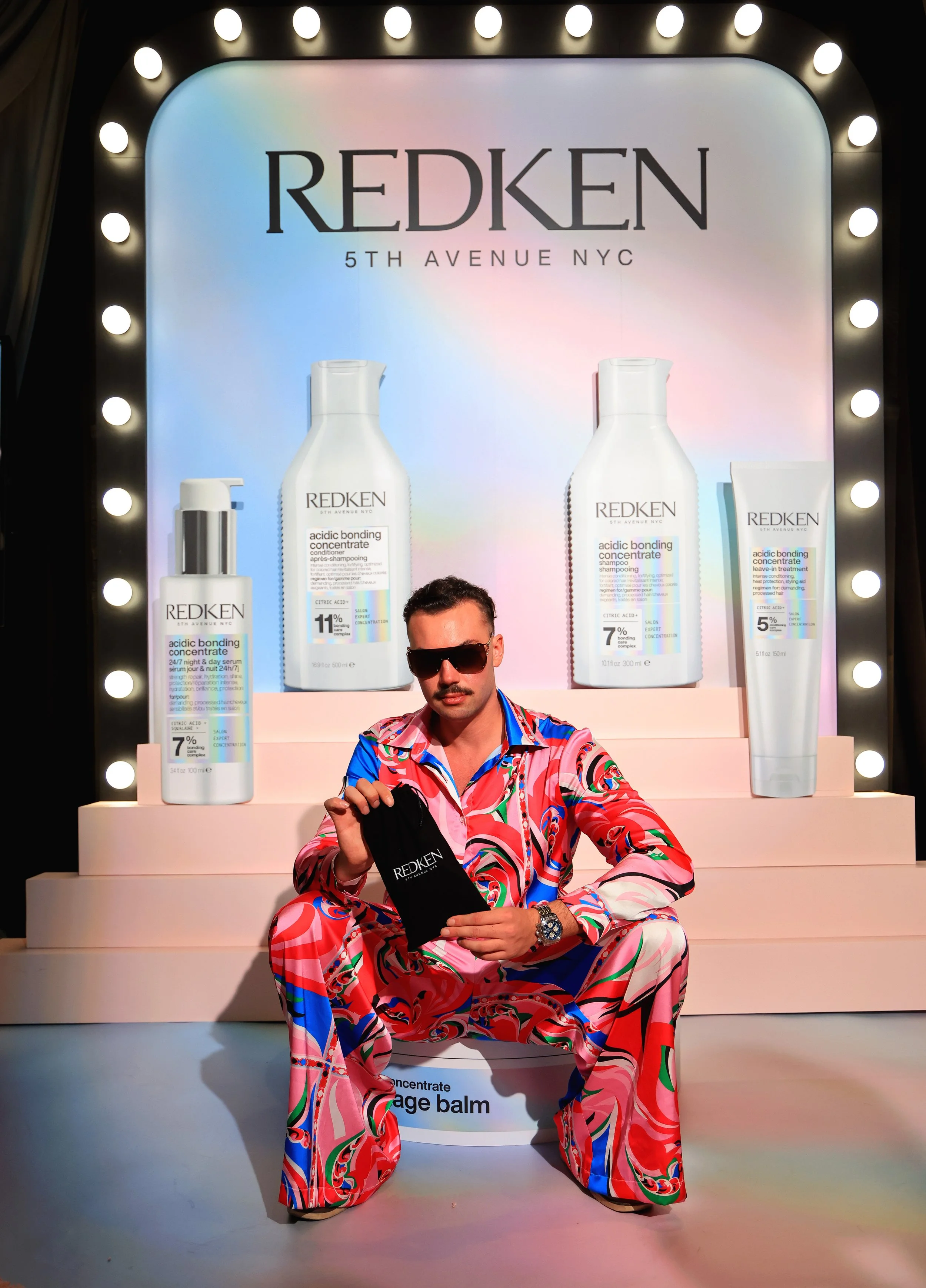 A man in colorful, patterned clothing sitting in front of a Redken hair care display, holding a small black pouch with Redken branding.