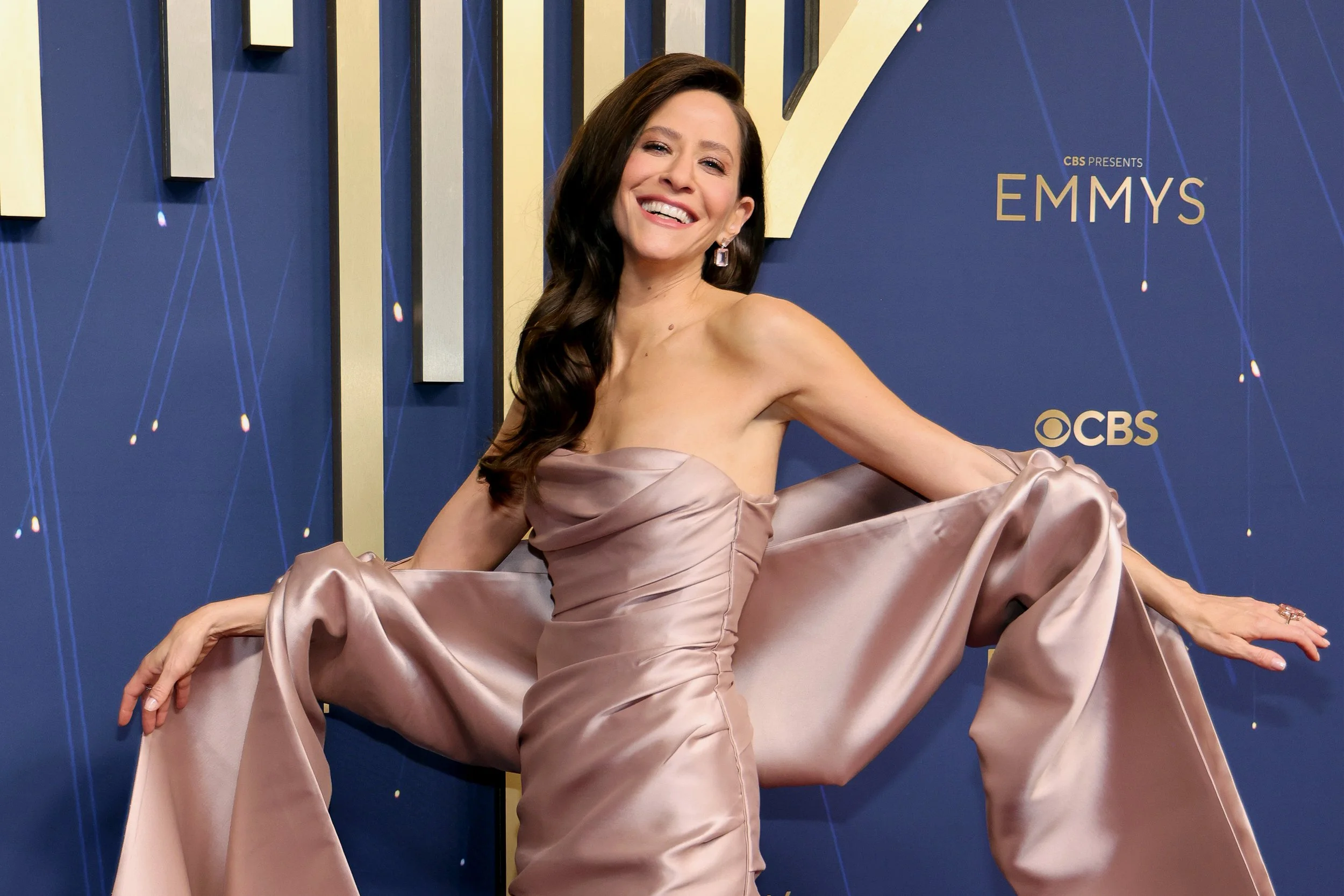 A woman in a strapless, satin gown with a dramatic train, posing on the red carpet at the Emmy Awards, smiling and stretching her arms outward.