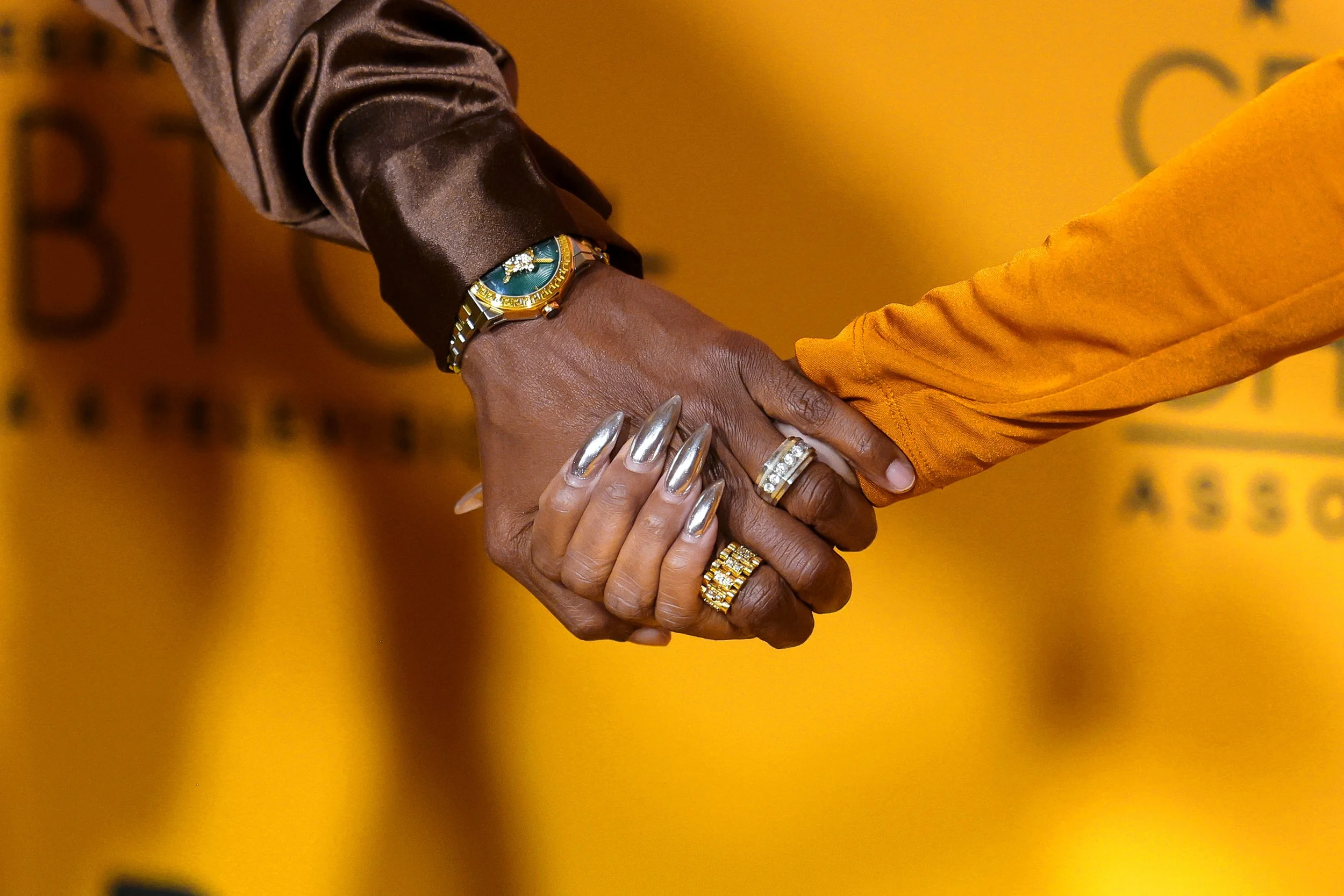 Two hands shaking, one hand with a bracelet and metallic nails, the other with rings, against a yellow background with blurred text.