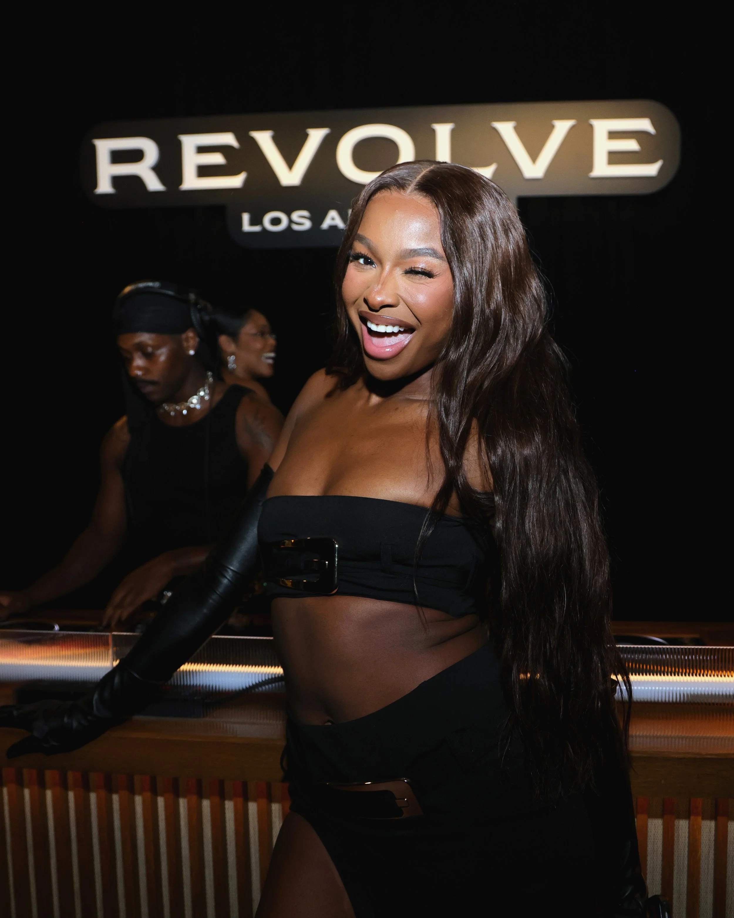 A woman with long brown hair, dressed in black, smiling and winking at the camera at a nightclub with a sign that says 'REVOLVE Los Angeles' in the background.