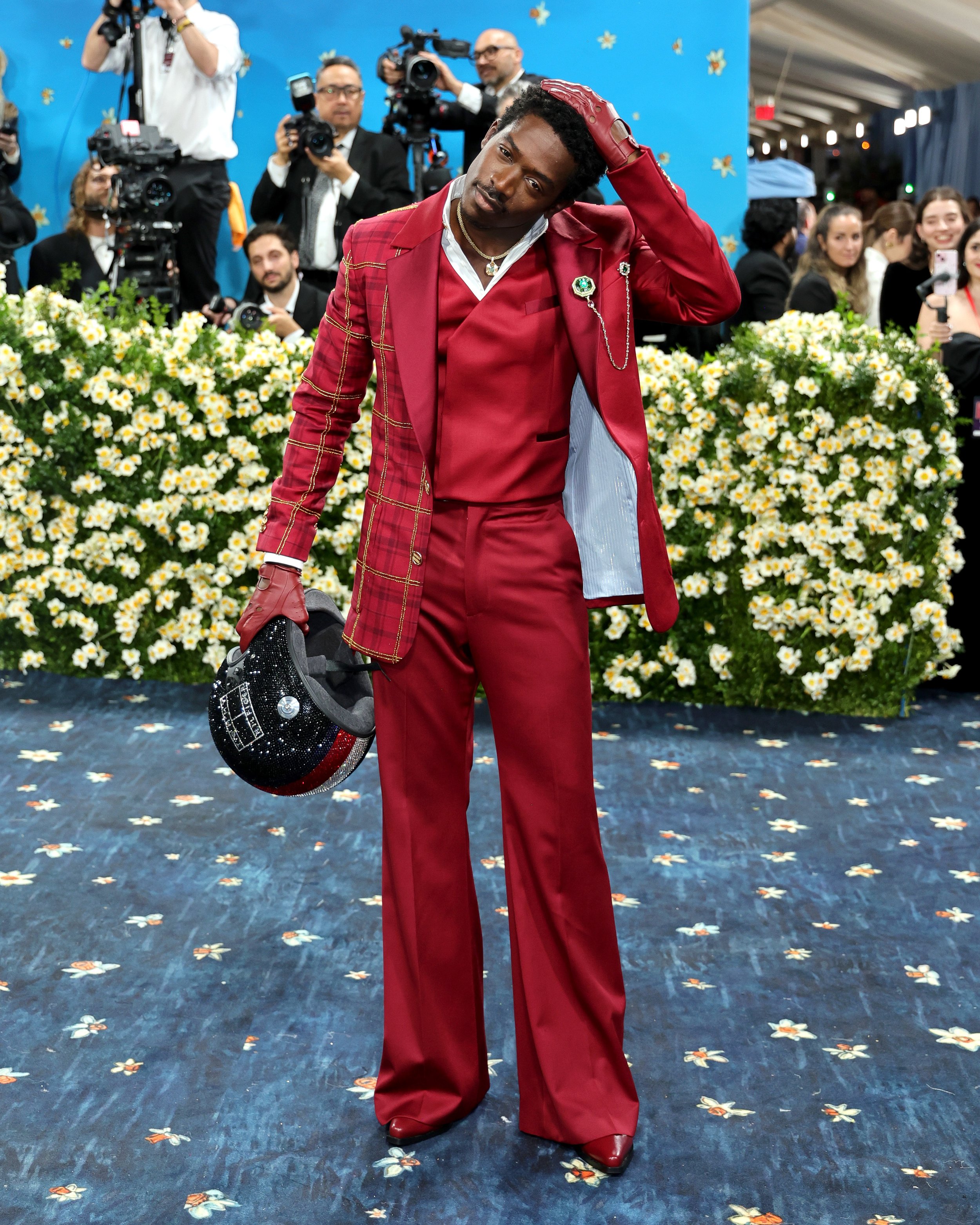 A man in a red suit holding a helmet, standing on a carpeted floor with a floral backdrop, surrounded by photographers and an audience.